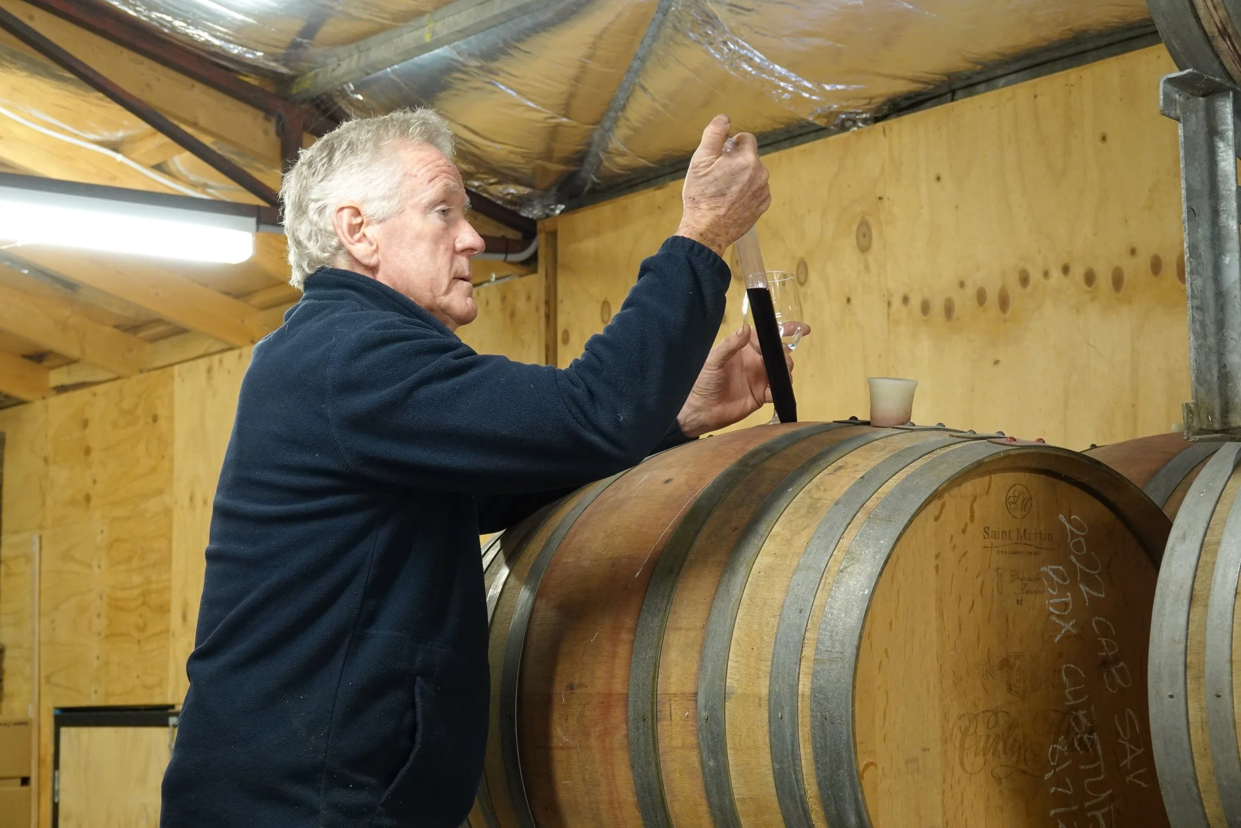 An elderly man with gray hair and a navy fleece examines a wine glass and a test tube of dark red wine in a winery cellar, with wooden wine barrels and wooden paneling in the background.