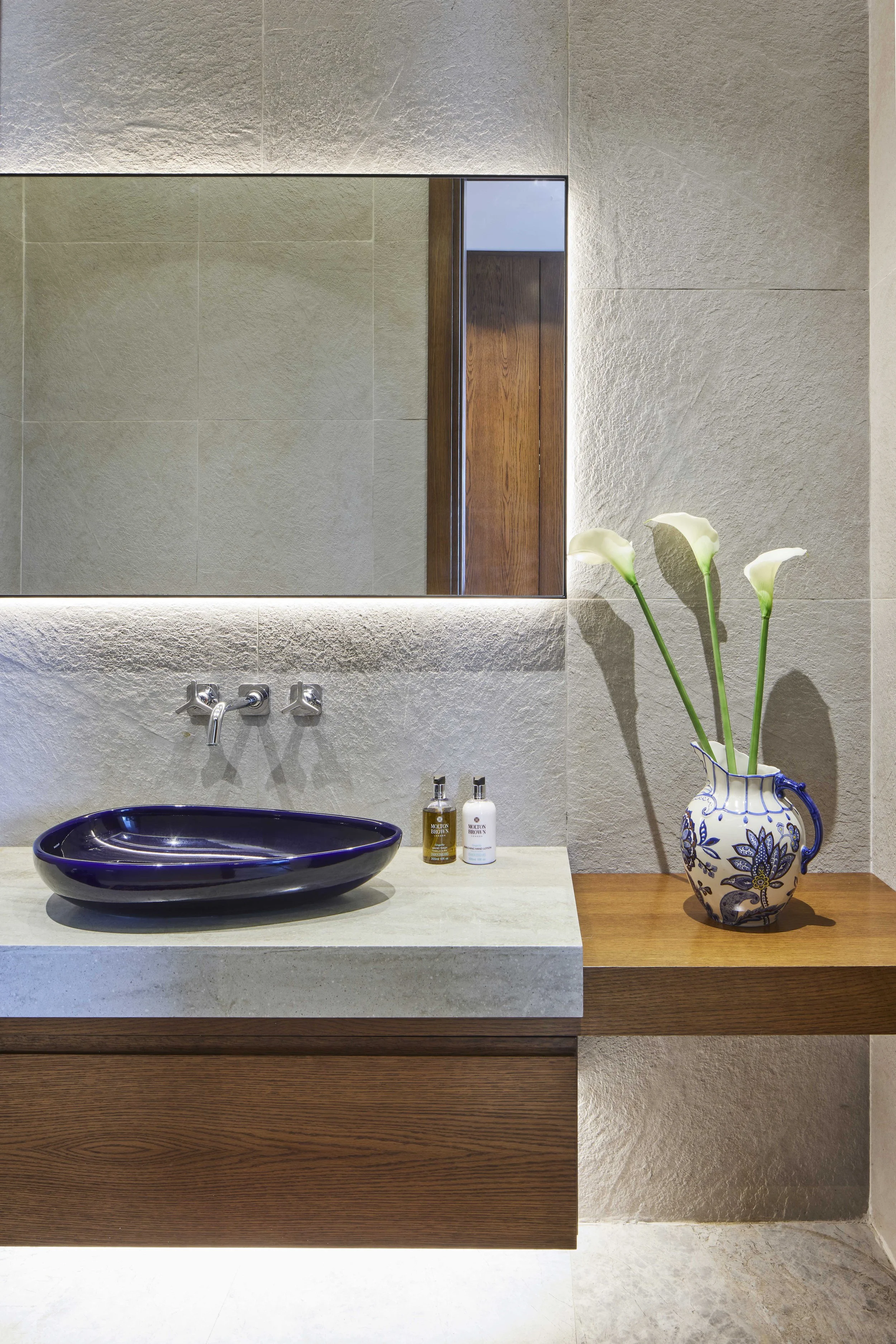 Modern bathroom sink with a purple bowl, a decorative vase with white calla lilies, and toiletry bottles on a light-colored countertop, with a large mirror and textured wall tiles.