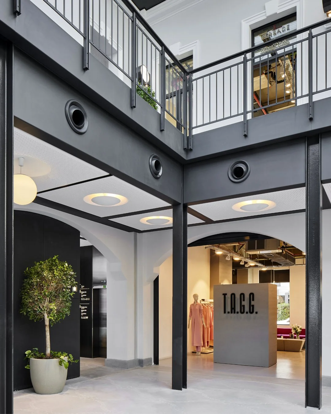 Interior view of a modern commercial building with a black and white color scheme, a second floor balcony with a black metal railing, a potted plant, and a clothing store named T.A.G.G. visible inside.