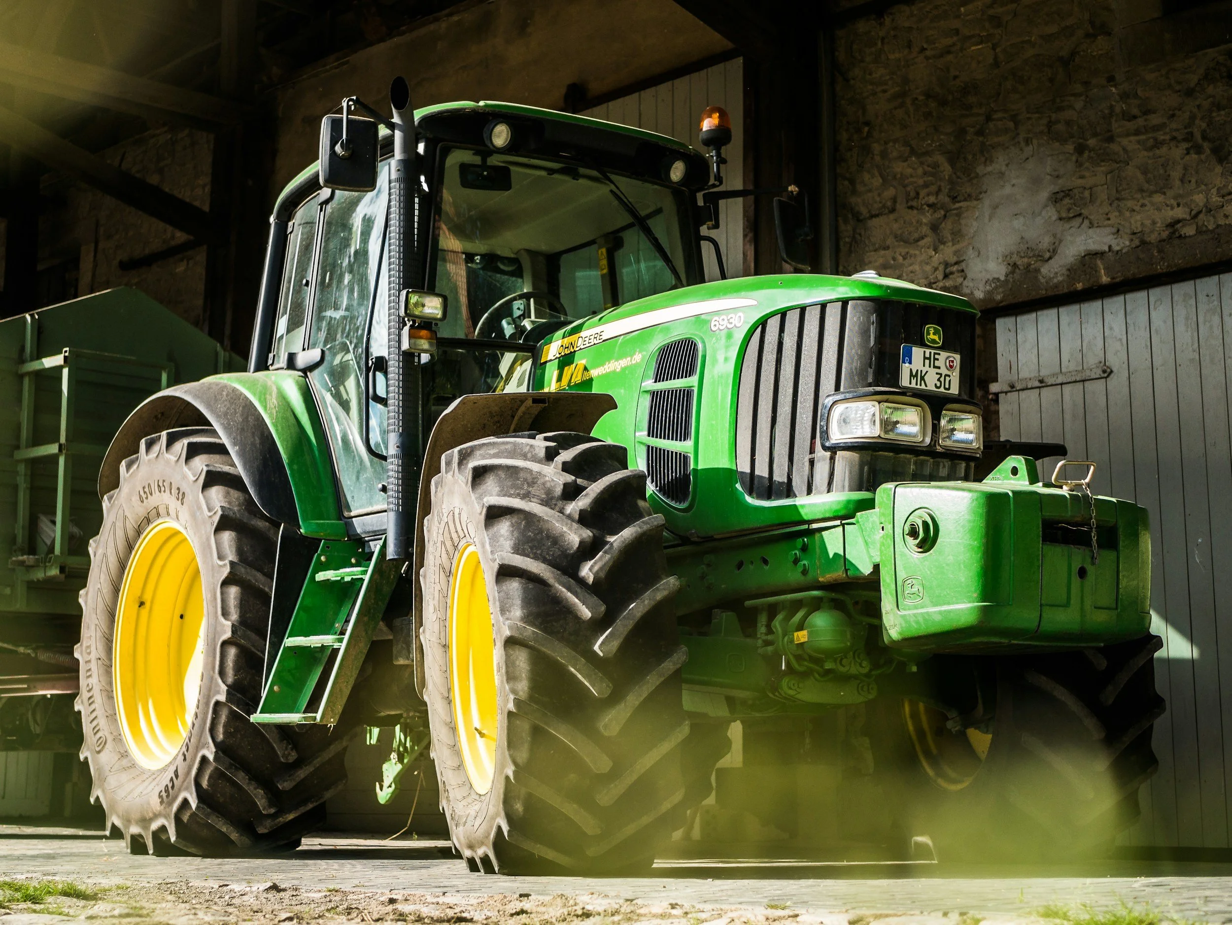 Green John Deere tractor parked indoors with large tires and a chain on the front, in a barn or garage setting.