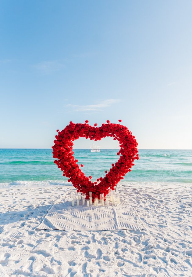 A heart-shaped floral arrangement made of red roses on the sandy beach with the ocean and sky in the background.