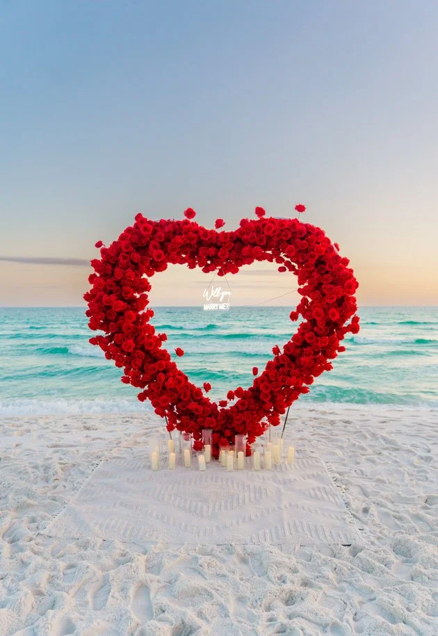 A large heart-shaped floral arrangement made of red roses on a beach at sunset, with candles placed at the base and a small sign that says 'Will you marry me?' hanging inside the heart.