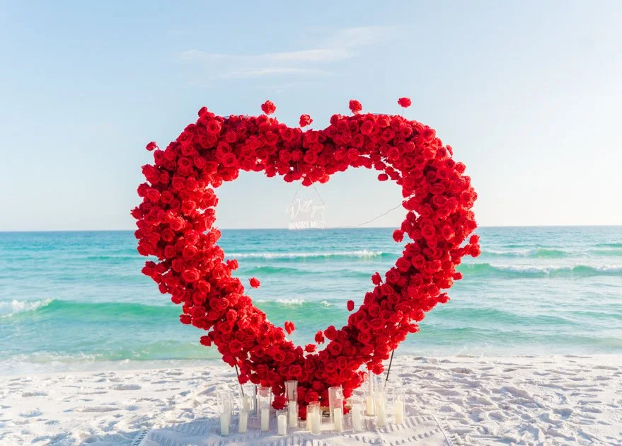 Heart-shaped arrangement of red roses on a beach with ocean waves in the background.