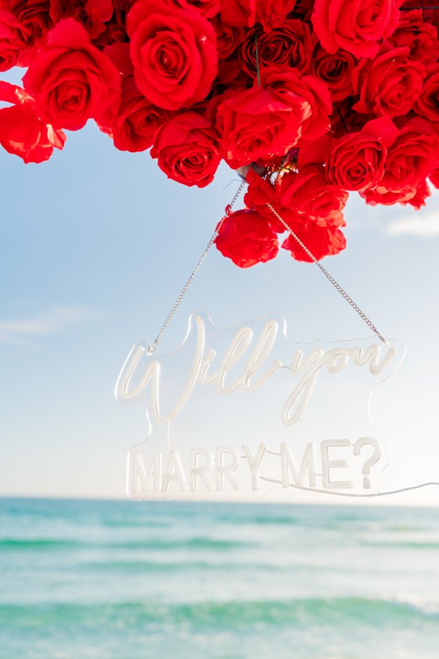 A white sign that says 'Will you marry me?' hanging from a bunch of red roses at the beach with ocean waves in the background.