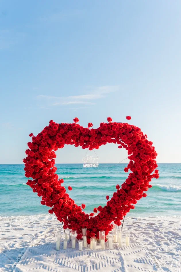 A large heart-shaped frame made of red roses on a beach with white sand and ocean waves in the background, with a small sign that says "Will You Marry Me?" hanging inside the heart.