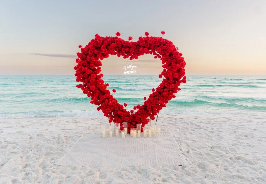 A large red heart made of roses on a beach with candles at the base and the ocean in the background.