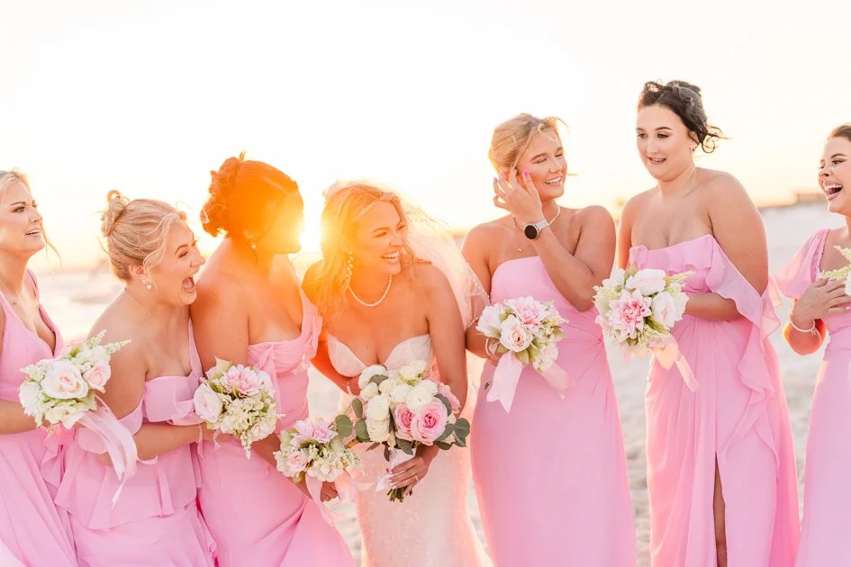 A group of women in pink dresses holding bouquets, smiling and laughing on a beach during sunset, celebrating a wedding.