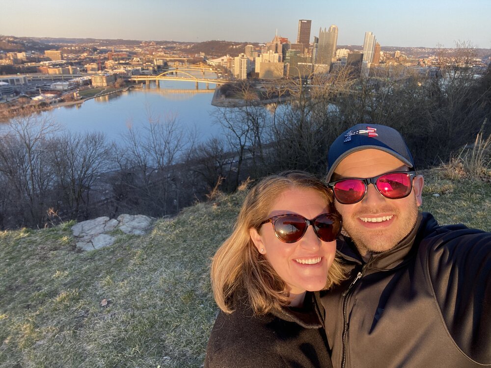 A couple poses for a selfie in front of the Pittsburgh skyline.