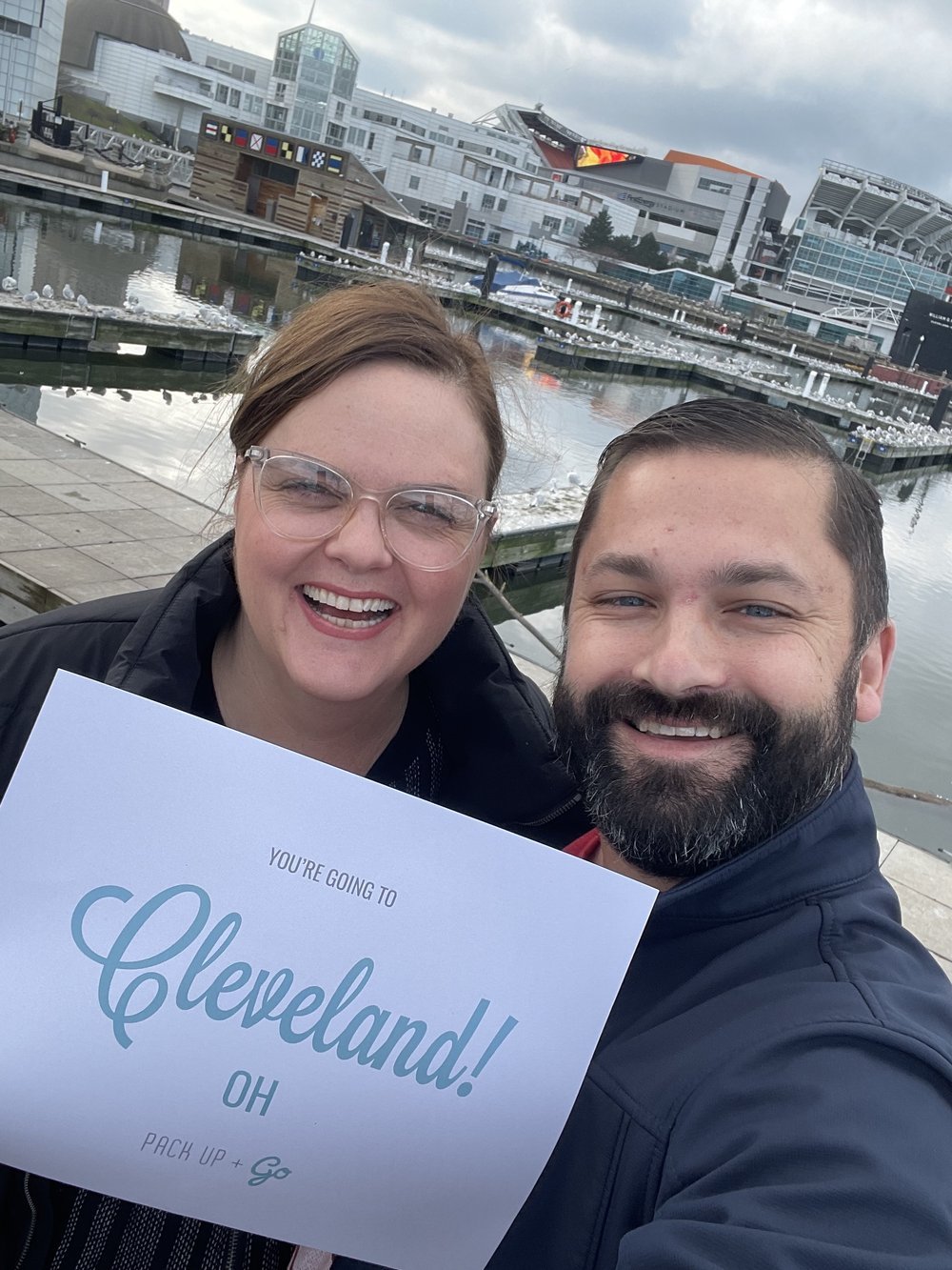A couple poses for a picture in a marina holding a destination sign saying, "Cleveland, OH"