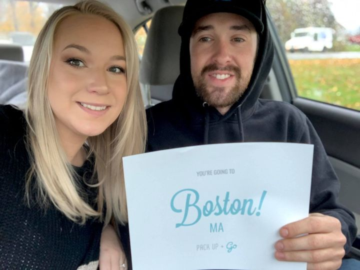A couple poses for a selfie in the car, holding a destination reveal sign saying, "Boston, MA!"