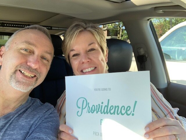 A couple poses in the car with a destination reveal sign saying, "Providence, RI!"