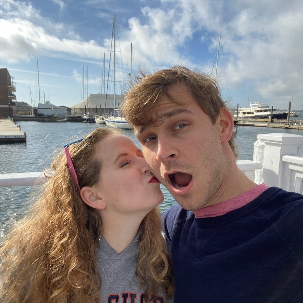 A woman kisses the cheek of a man in a marina.