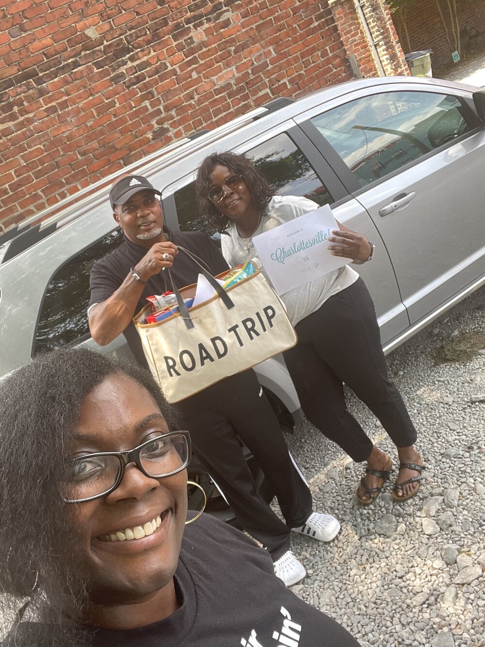 A family poses for a selfie in front of a car. One traveler is holding a destination reveal sign that says, "Charlottesville, VA" and the other is holding a tote bag with the words "Road Trip" printed on the front.