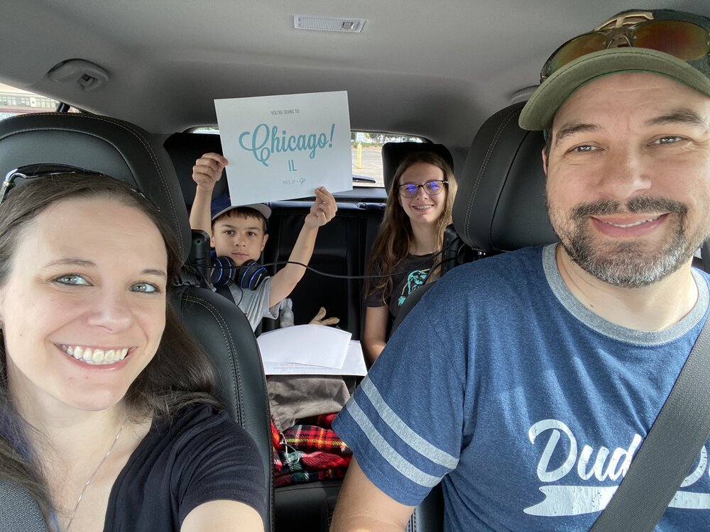 A family poses for a car selfie, and the child in the back holds a destination reveal sign saying, "Chicago, IL!"