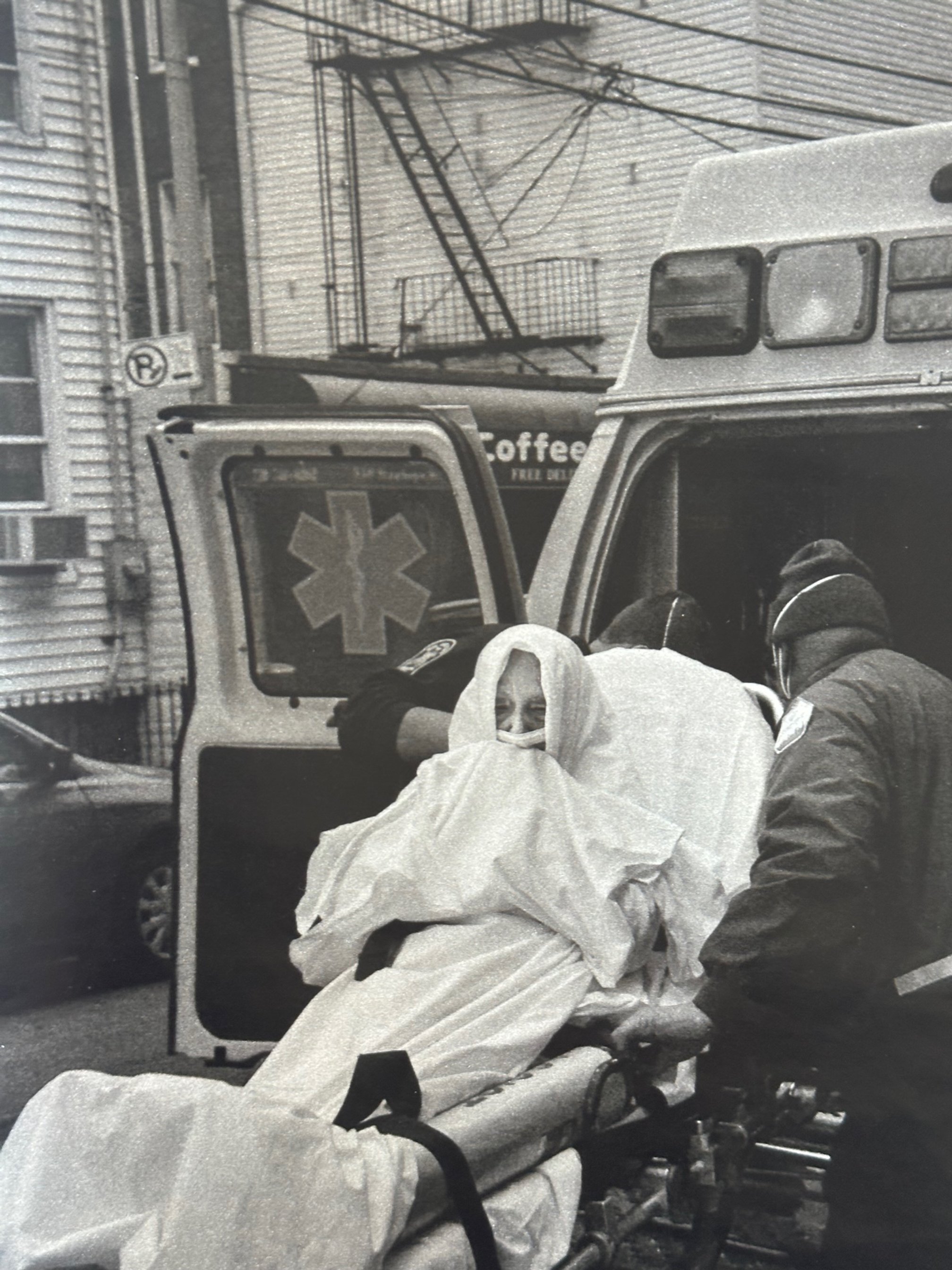  Black and white image of person on stretcher with mask on being loaded into ambulance. 
