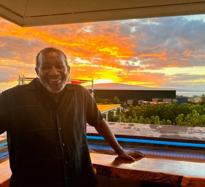 Bryant Neal with gray hair and beard smiling, standing against a balcony at sunset, with a view of clouds and trees in the background.