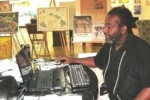 A man with a beard working at a desk with a laptop, computer monitor, and keyboard in an office or workspace.