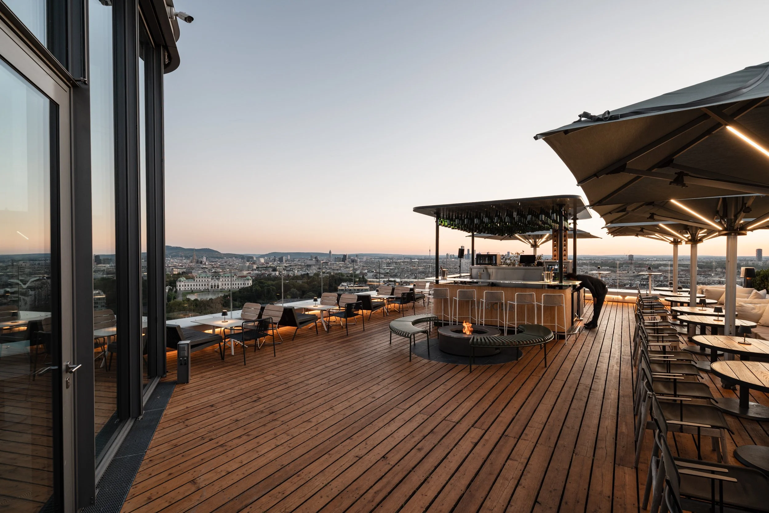 Dachterrasse mit Bar, Sitzplätzen und Feuerstelle bei Sonnenuntergang mit Blick über Wien und das Schloss Belvedere