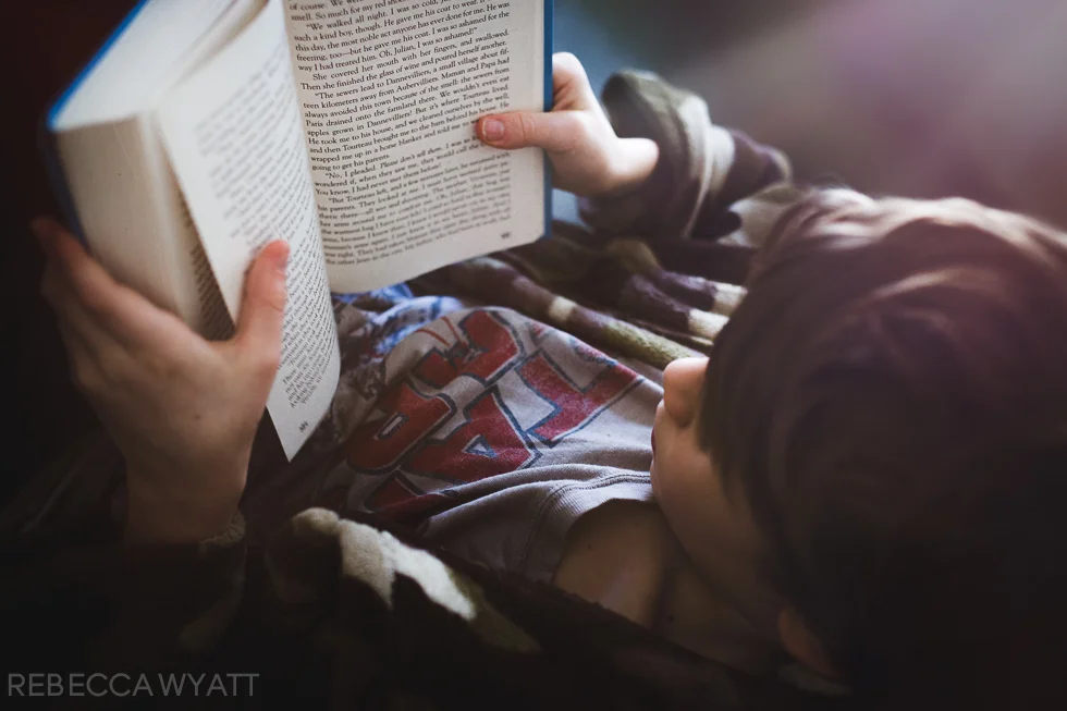 Boy in star wars shirt reading a book by a window.