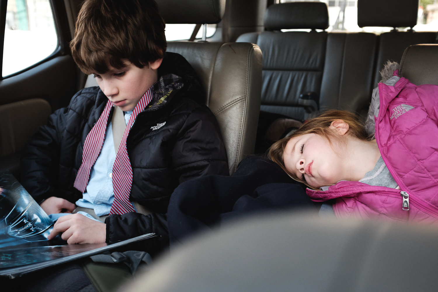 Boy reading in backseat of car with little sister sleeping beside him.