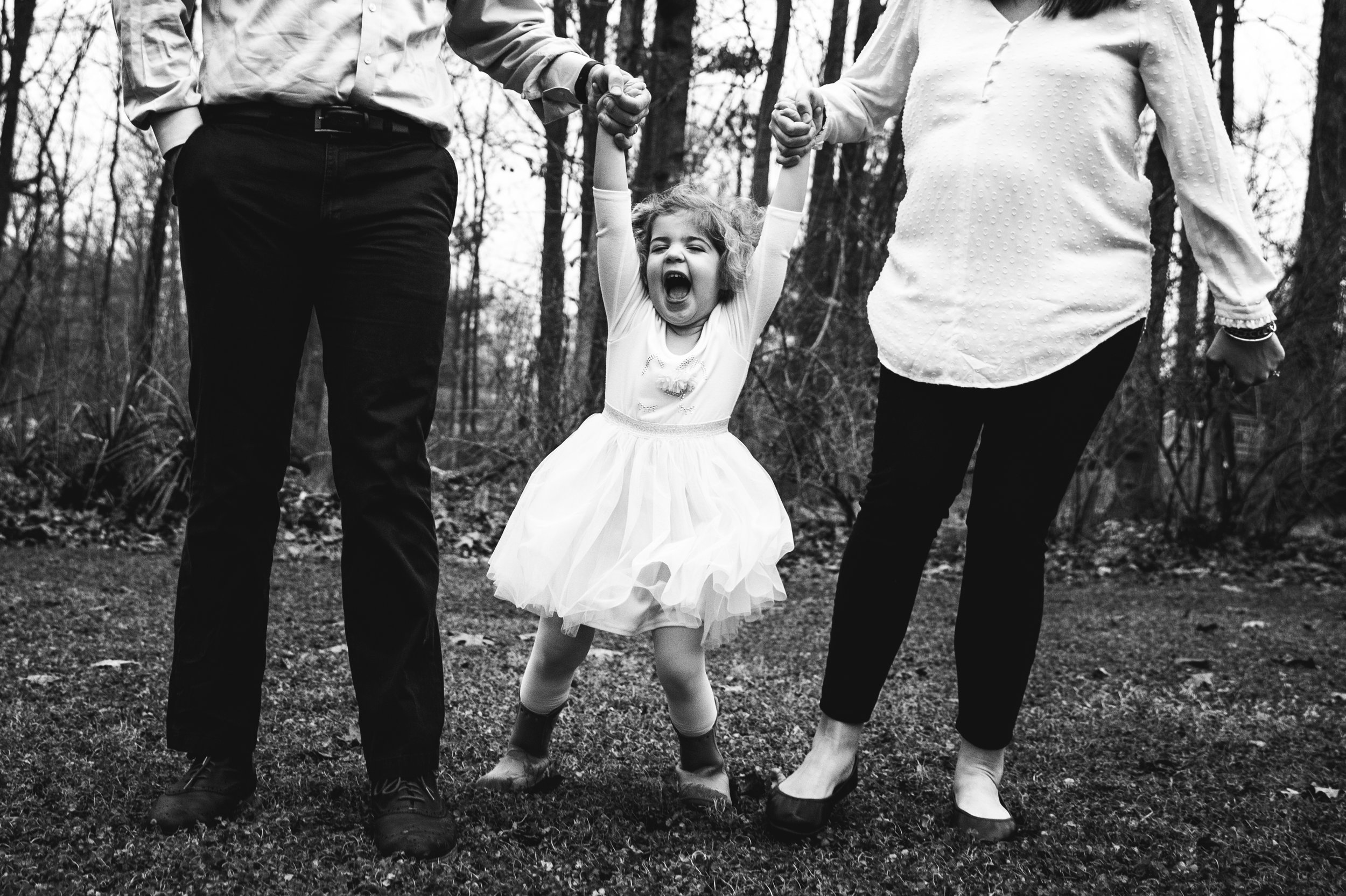 little girl in tutu hold's parents hands with big smile in black and white photograph