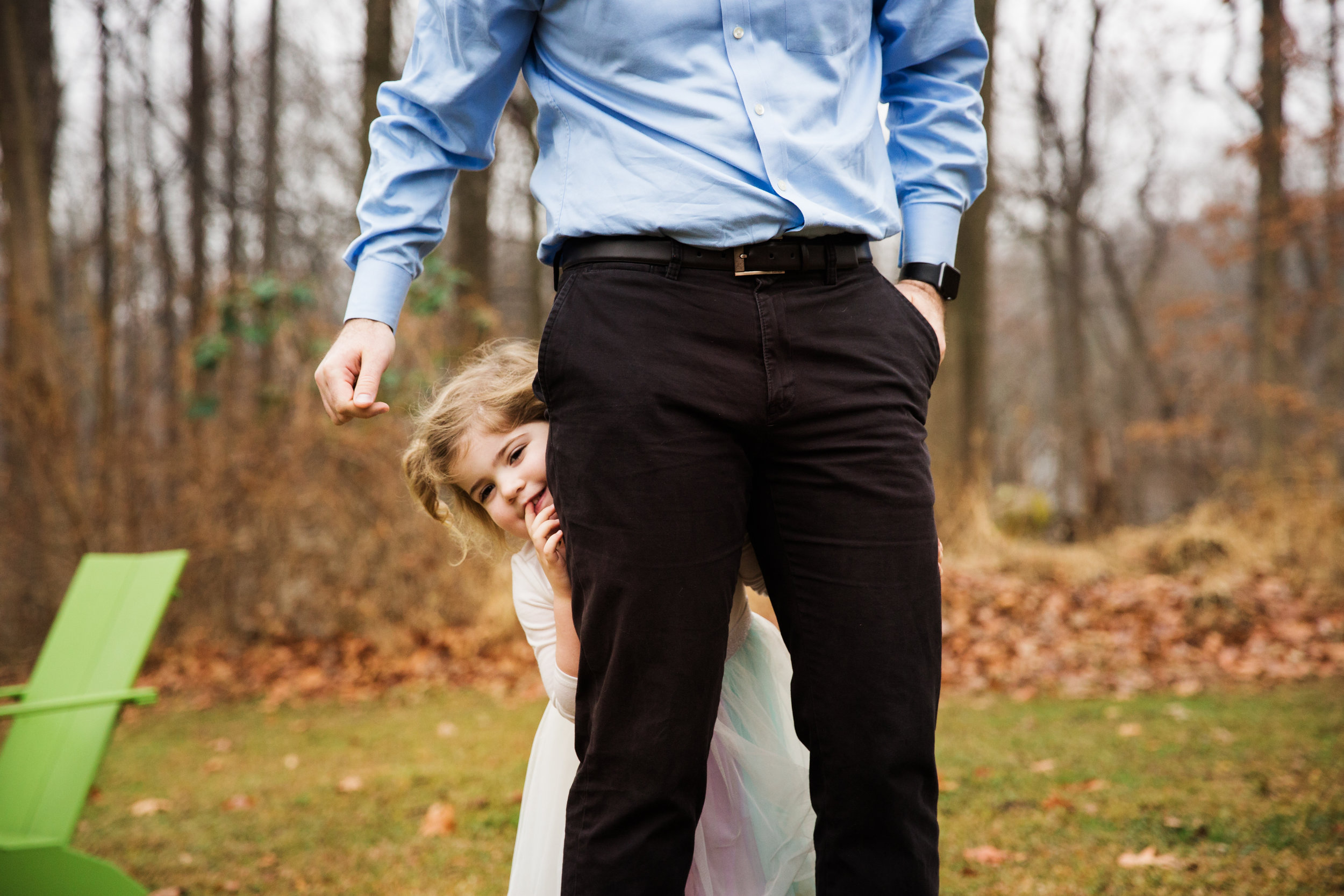 two year old girl peeks out from behind her dad's legs