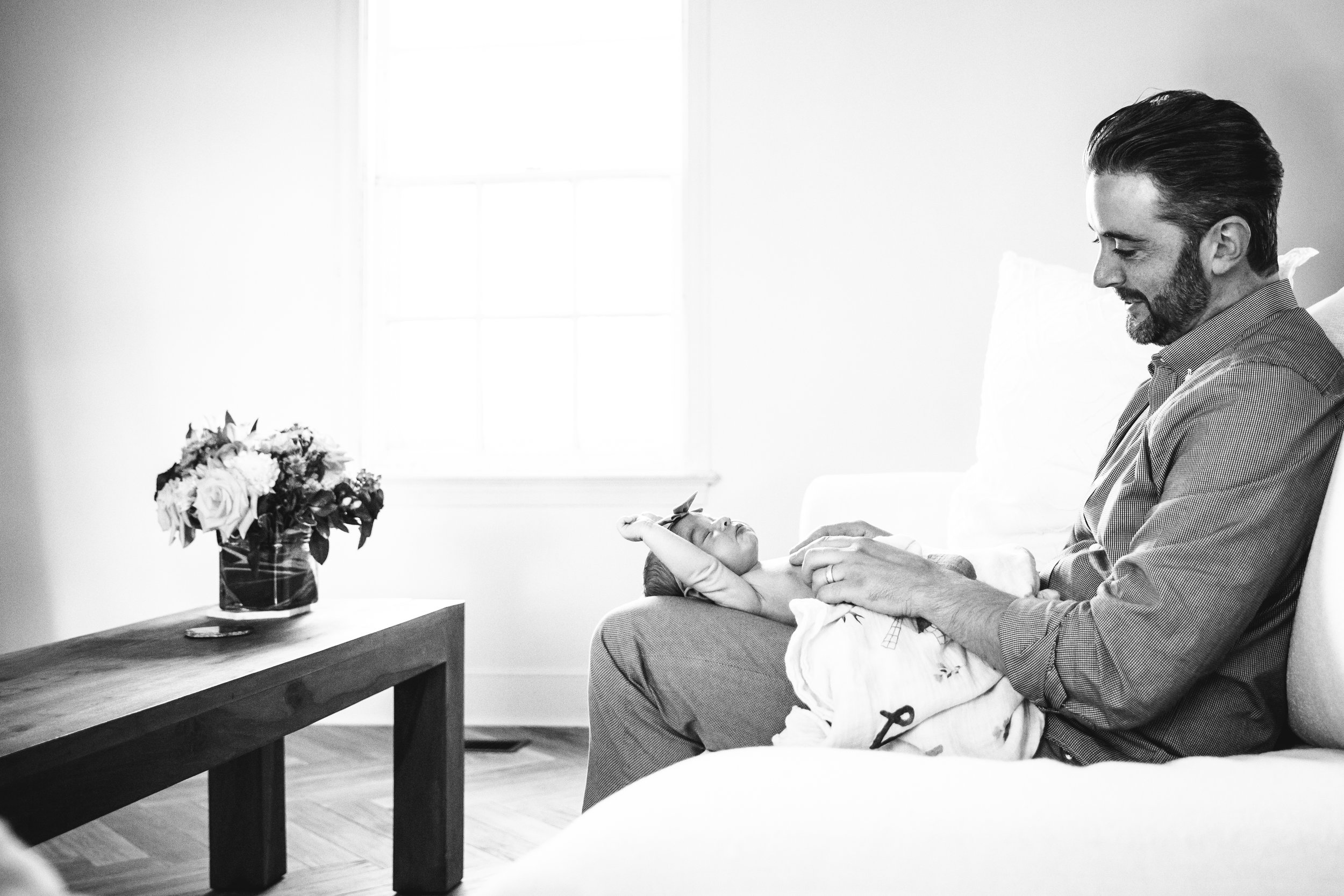 dad plays with his new baby daughter while sitting on sofa by a window 