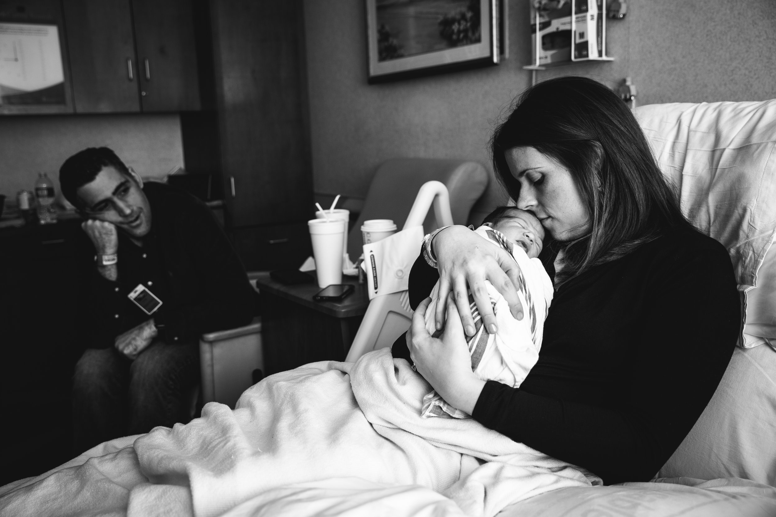 new mother snuggles and kisses infant son in hospital while baby's father looks on in background.