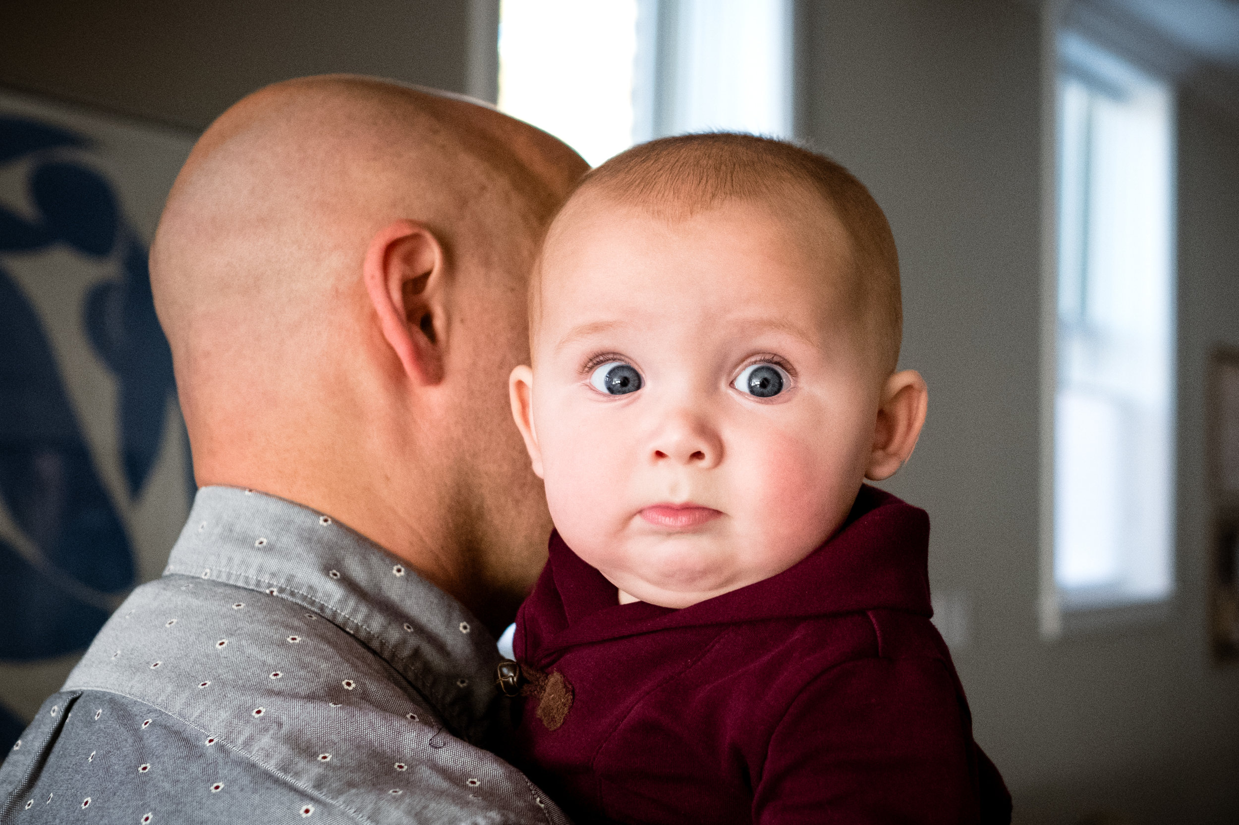 baby son in maroon shirt peeking over dad's shoulder looking at camera in Baltimore City home