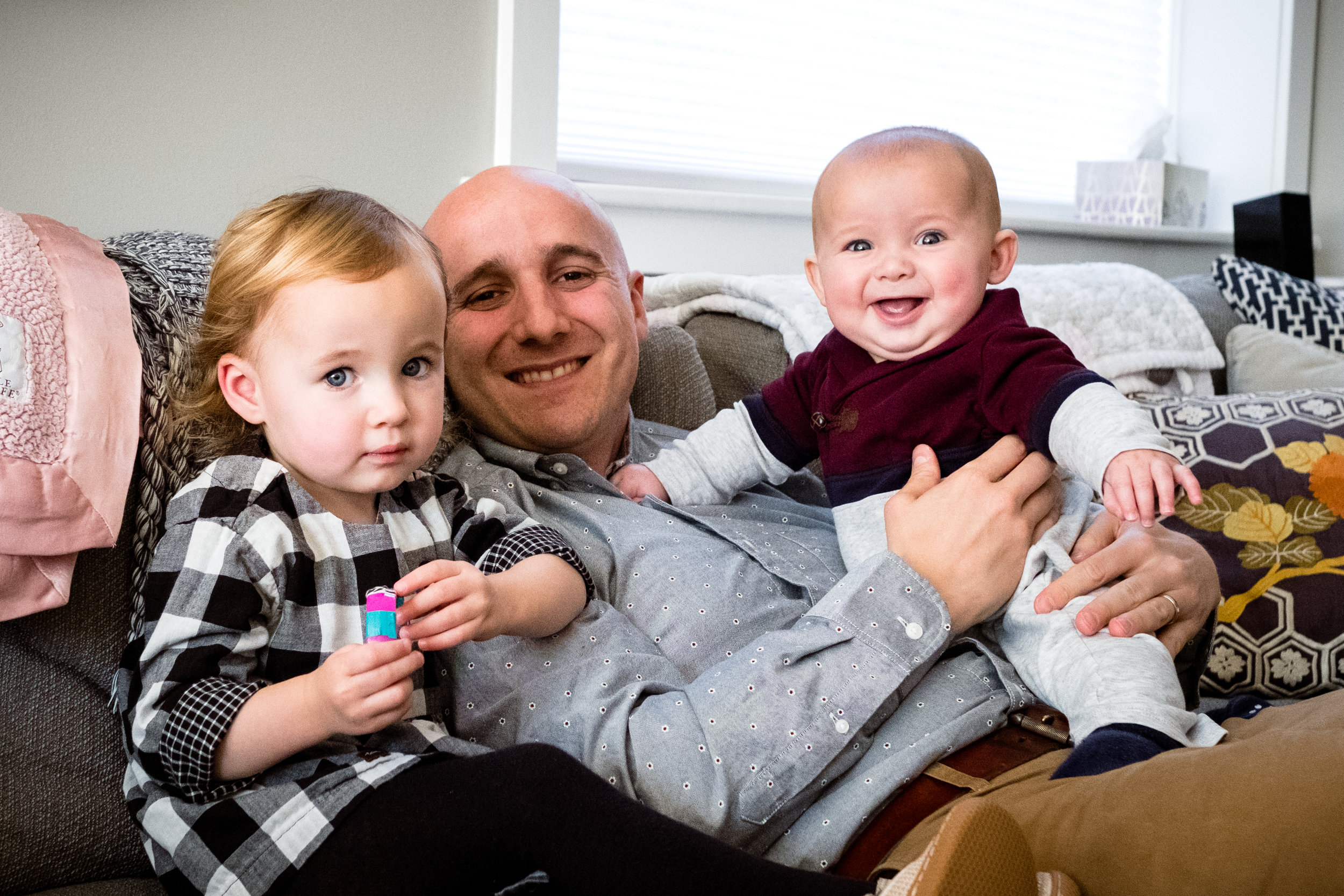 Father with young daughter and infant son smiling on sofa in their home.