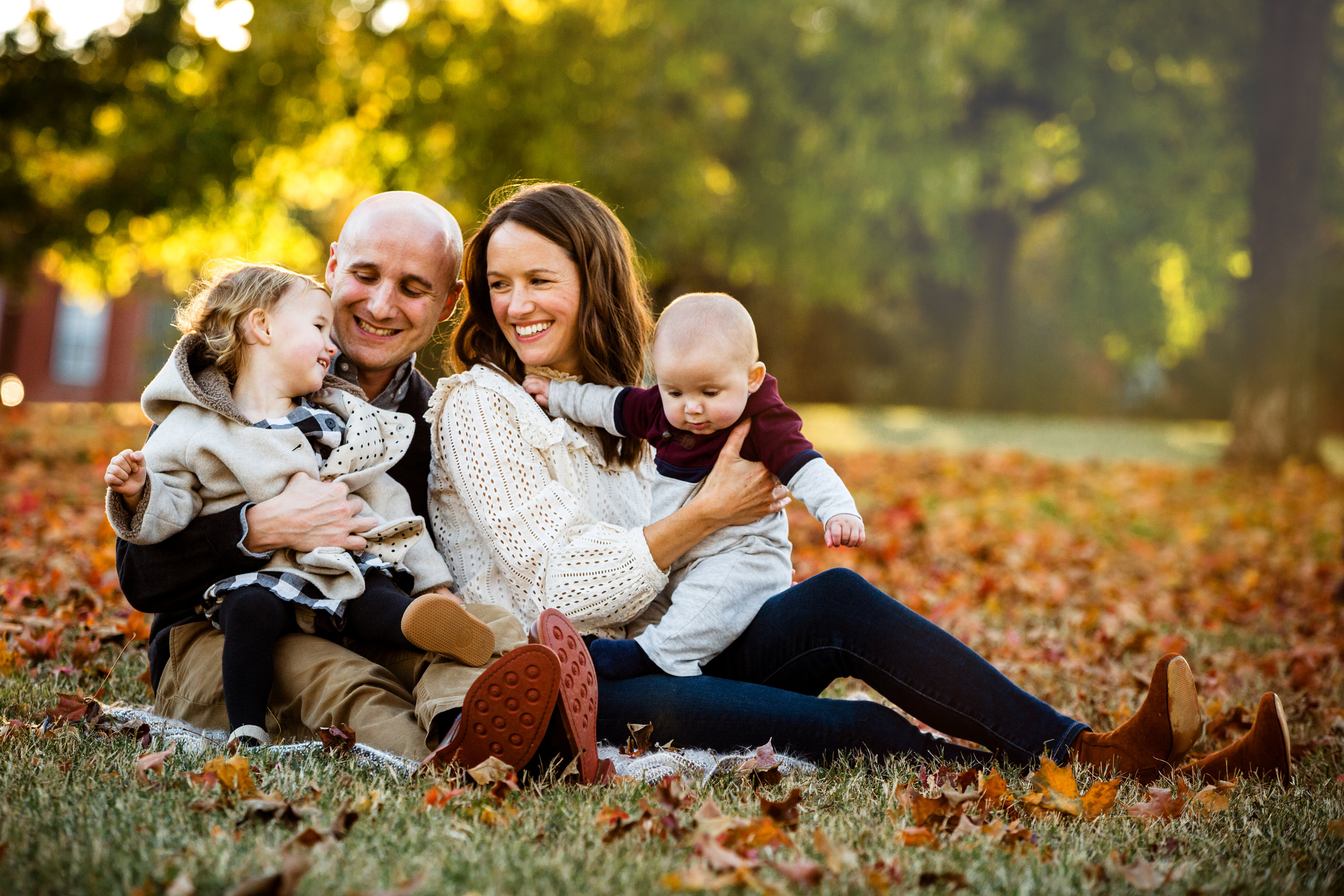 family of four, including dad, mom, daughter, and son, smile at each other while seated on grass amidst leaves