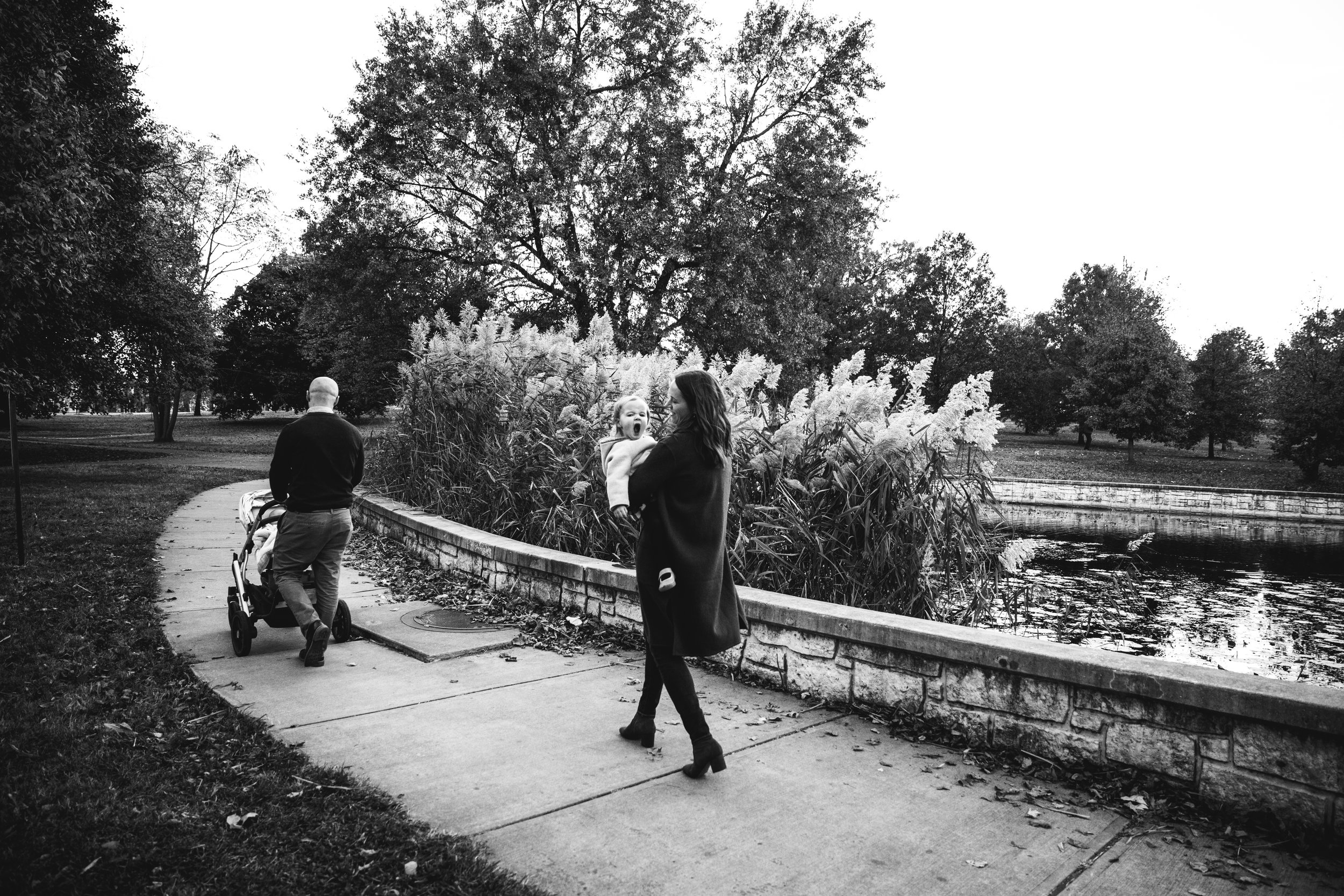 black and white photograph of family walking with stroller by park pond while mom holds daughter who yawns