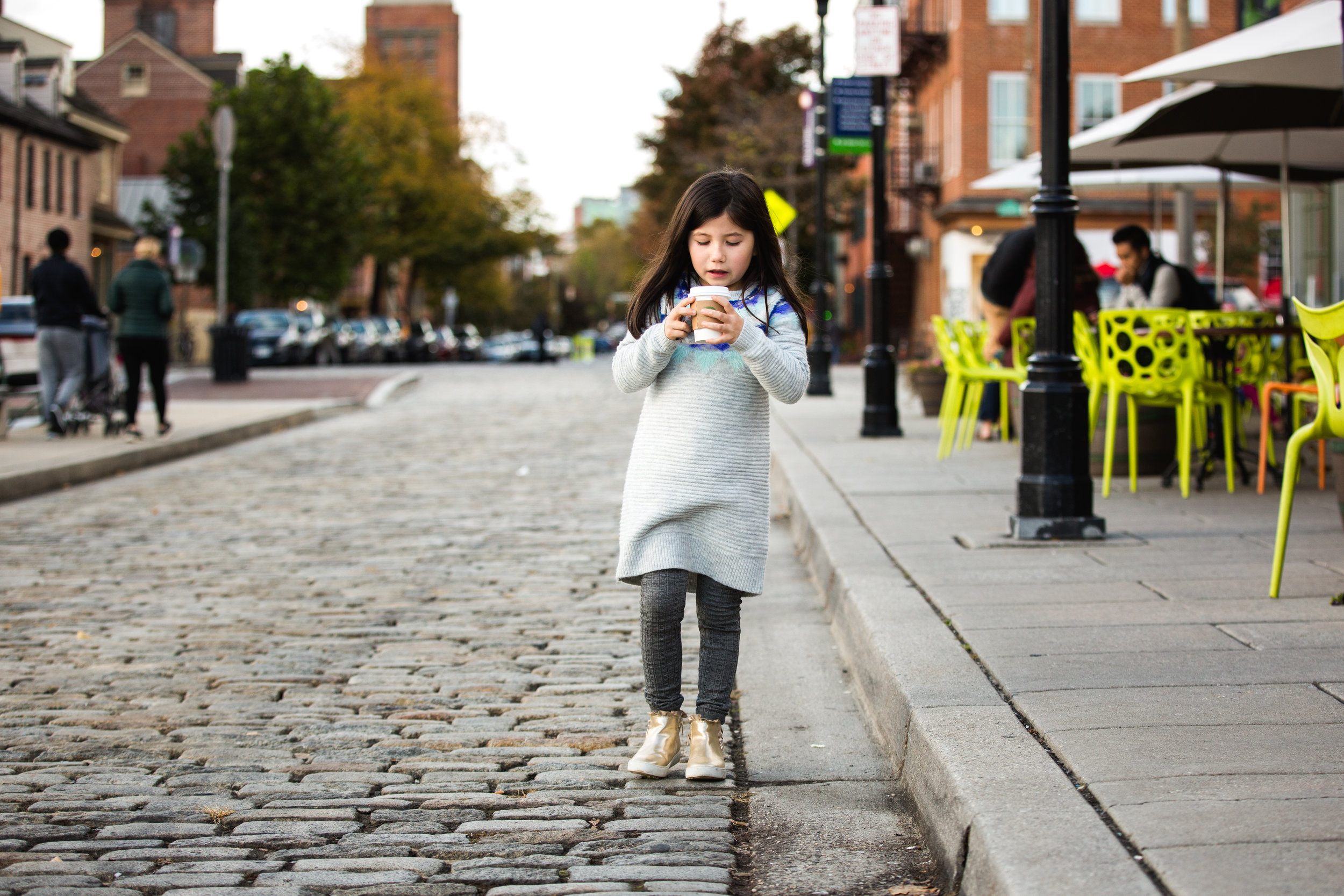 little girl in grey dress and leggings drinks hot chocolate on cobblestone streets of Fells Point
