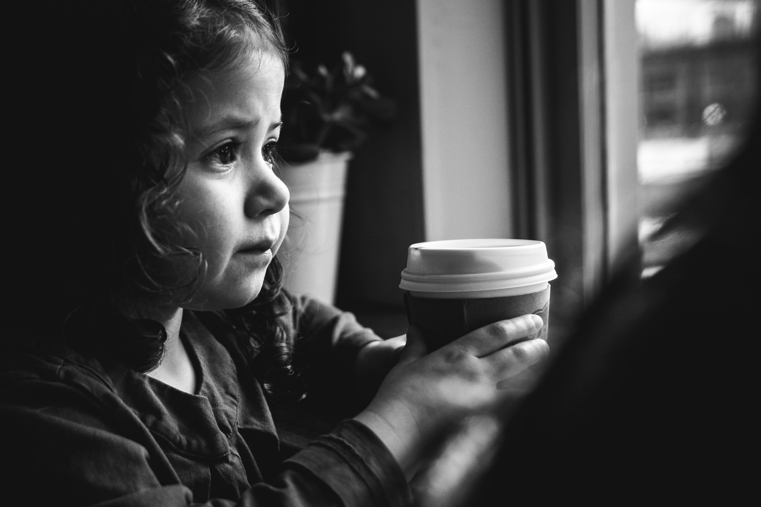 black and white photograph of toddler girl with dark curls looking out cafe window holding hot chocolate 