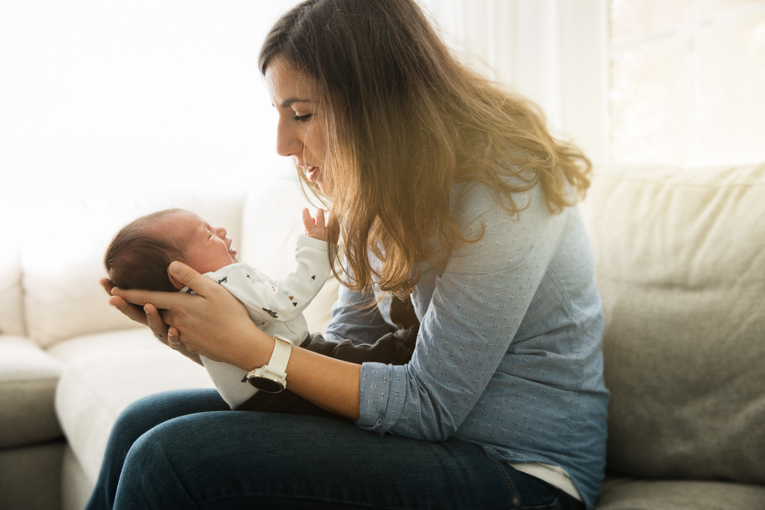 beautiful young mother leans in to look at baby daughter in backlit lifestyle newborn photograph