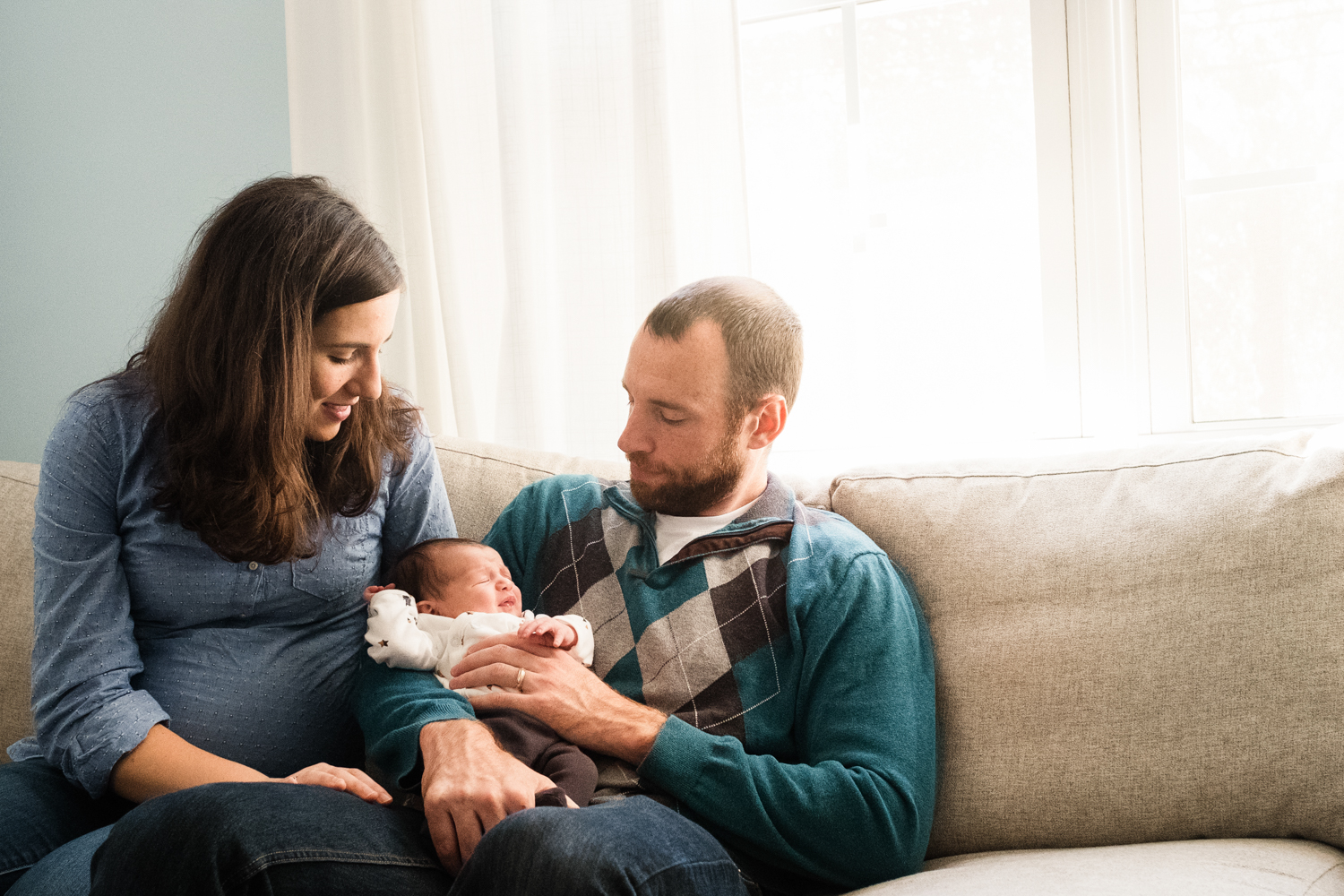 family of three including new baby sitting in family home on sofa by window