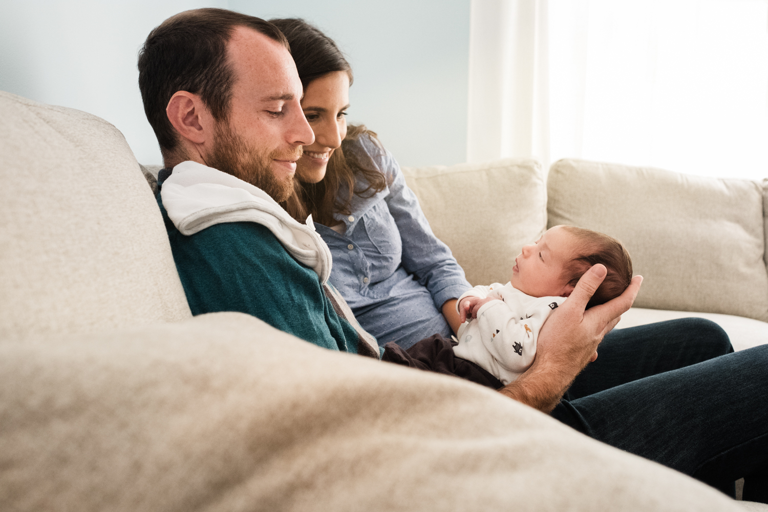 new parents admire their infant daughter while seated together on family sofa