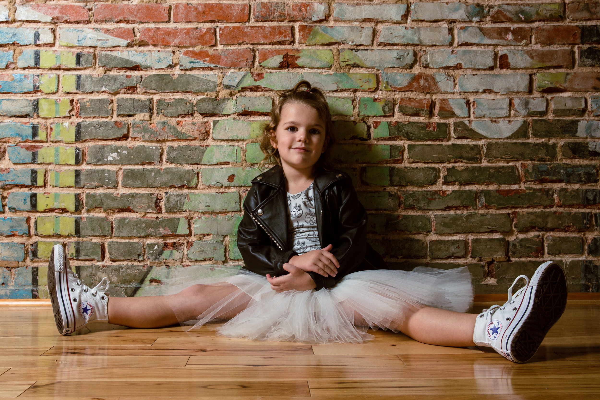 young child in leather jacket and tutu with converse sneakers sits against painted brick wall for portrait.