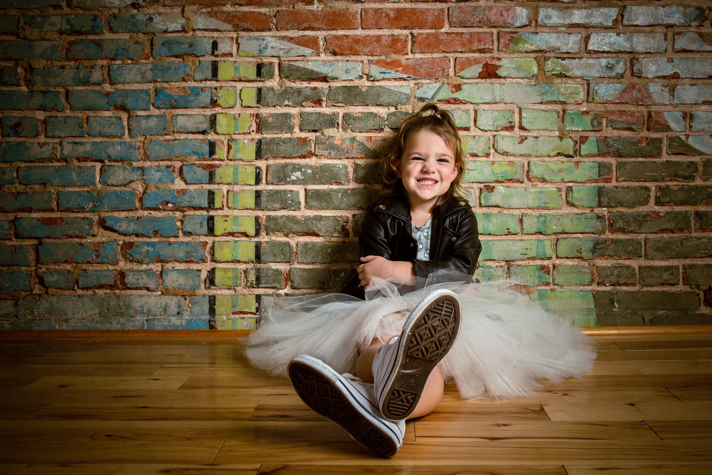 smiling little girl sits against brick wall wearing skirt and jacket, with sneakers on feet and legs crossed.