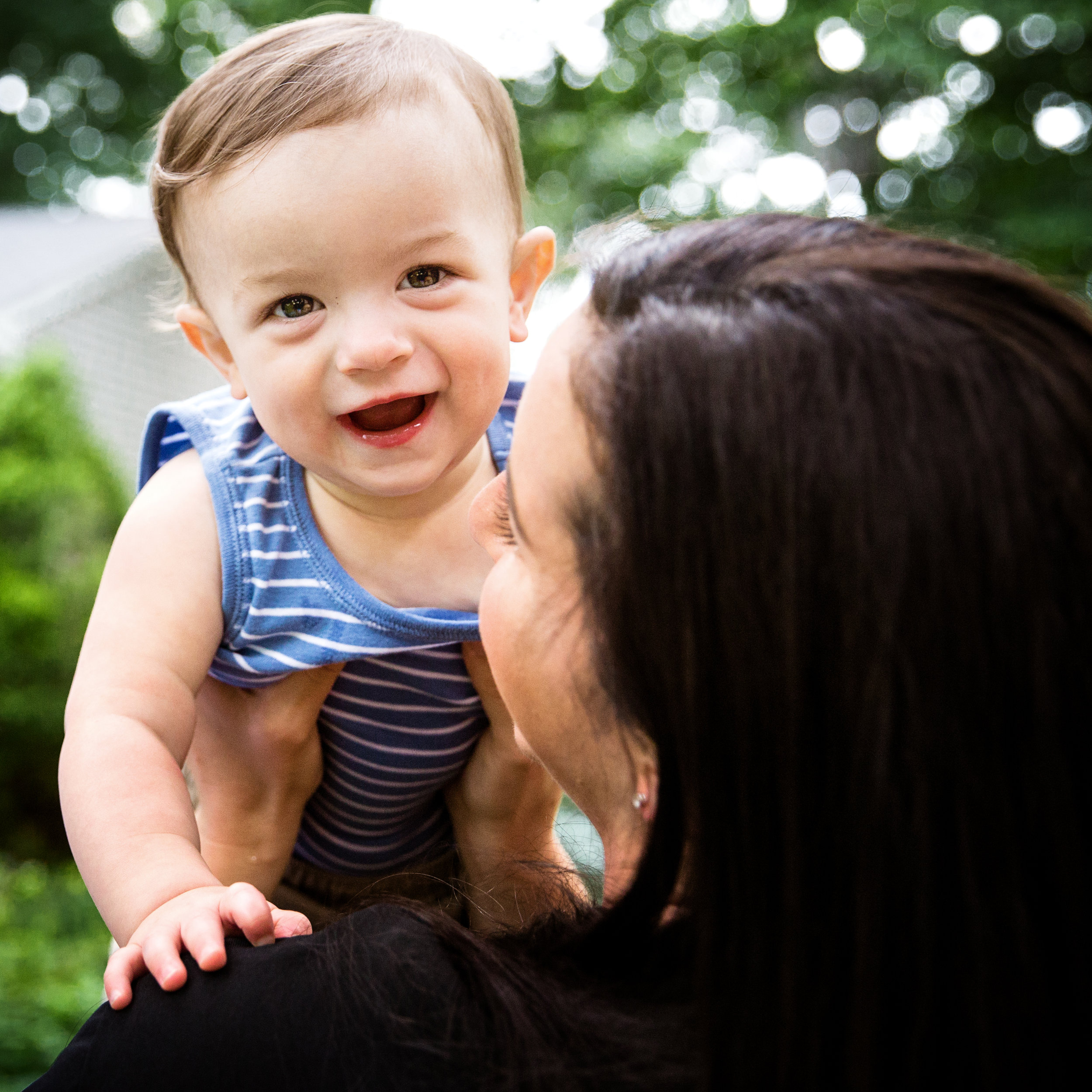 one year old boy peeks at camera for photographer over moms shoulder