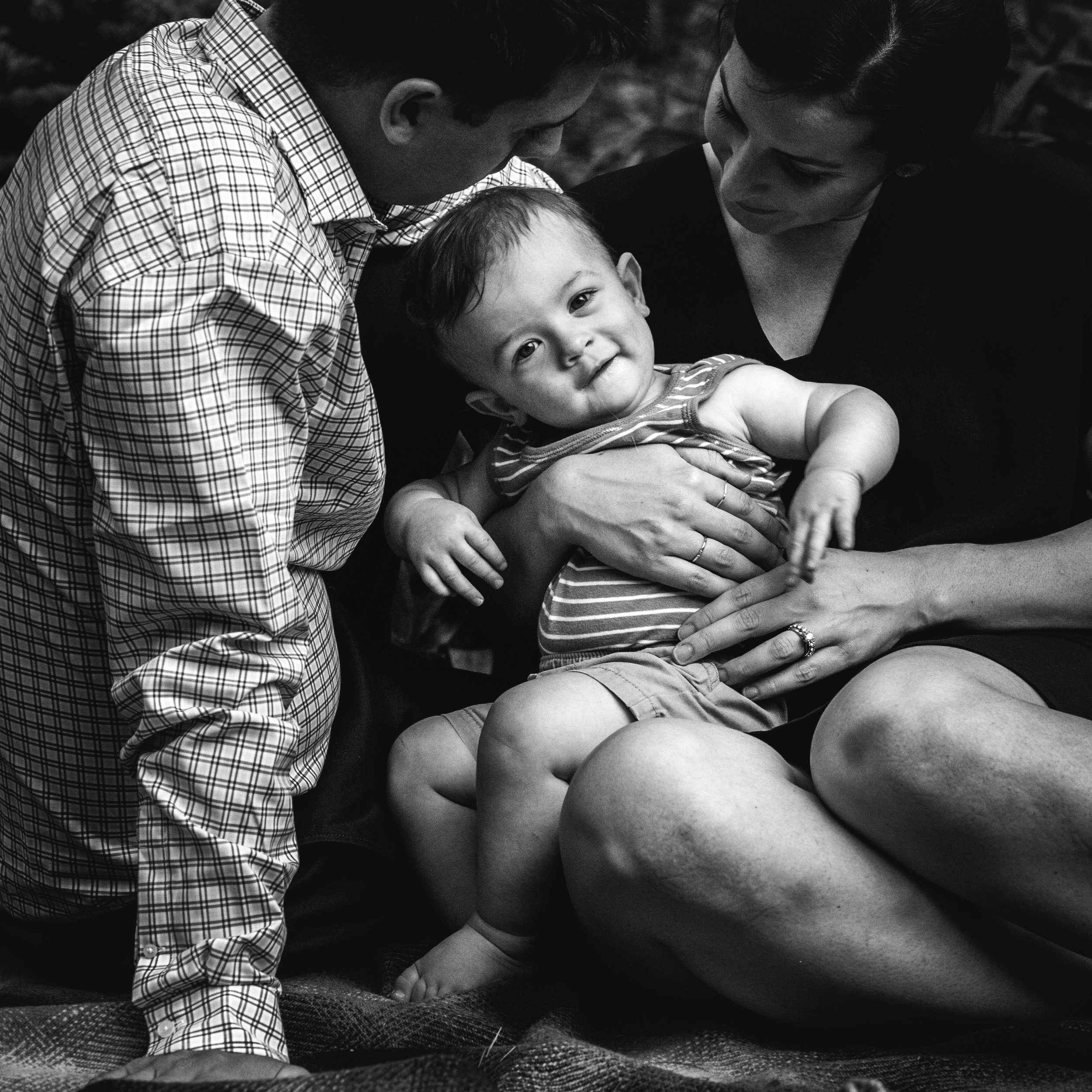 black and white photograph of smiling boy looking at camera while sitting in moms lap with dad near by