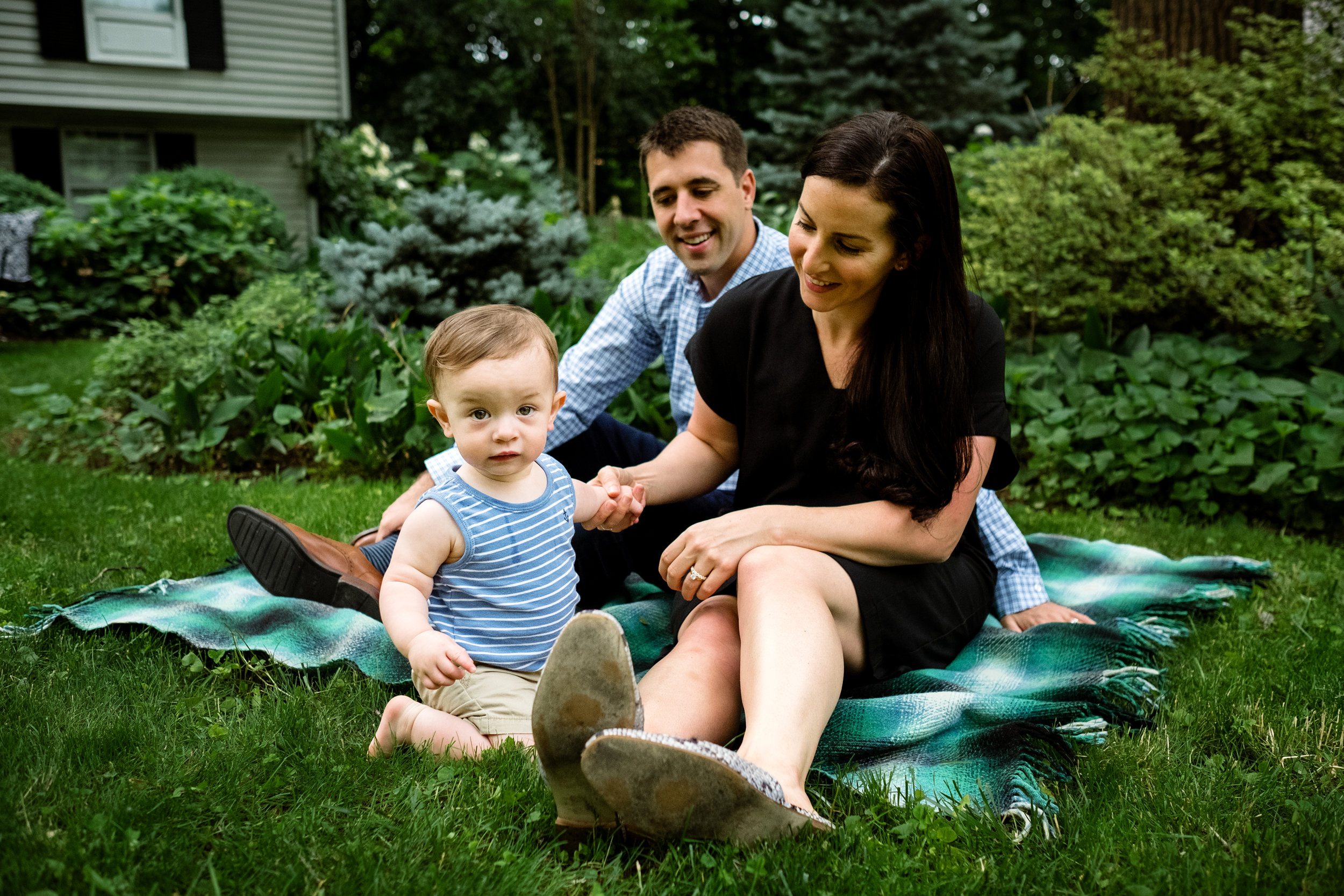 young family including parents and one year old boy sit on blanket in the grass