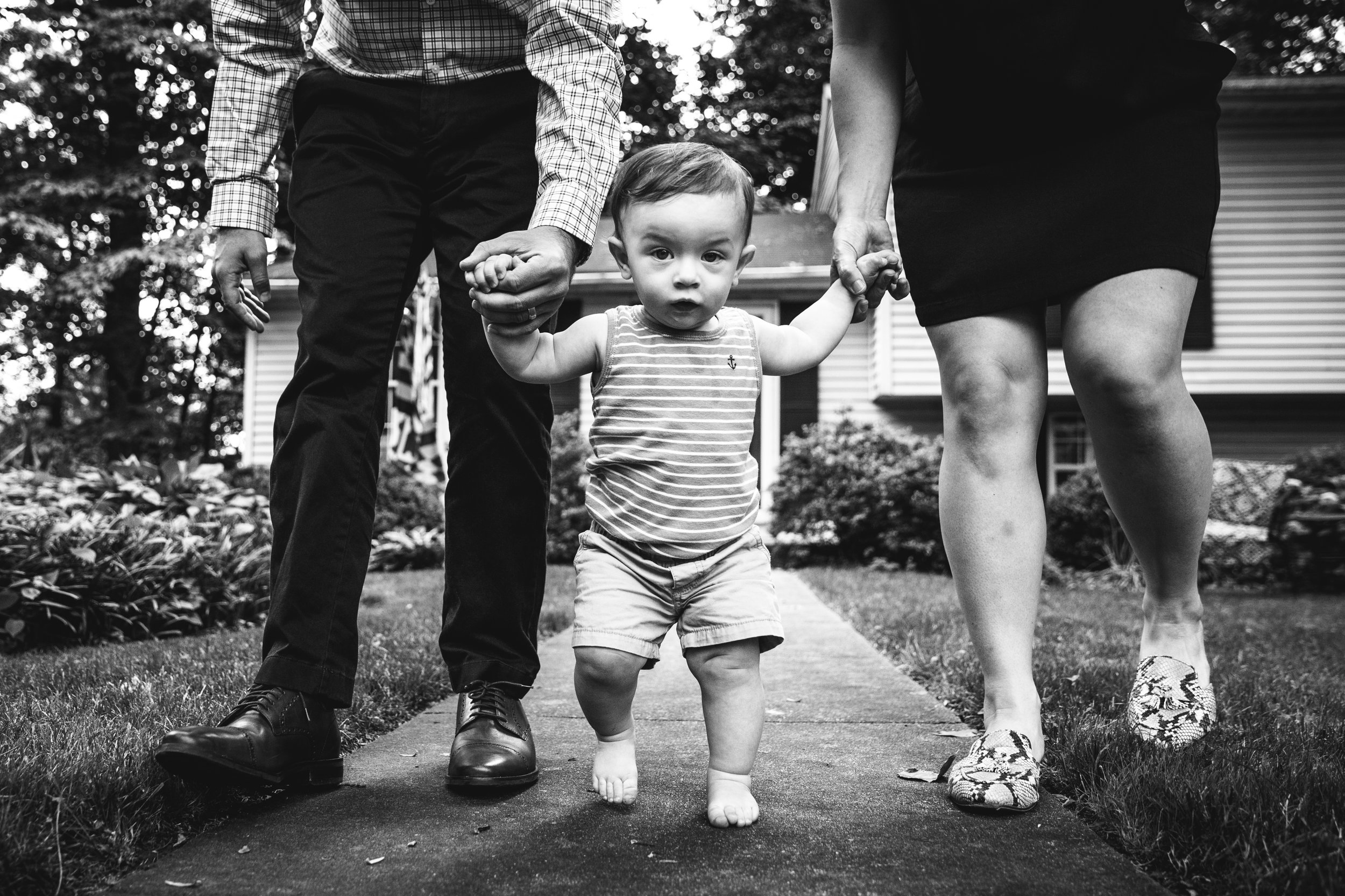 black and white photograph of little boy toddling on sidewalk being supported by parents holding his hands.