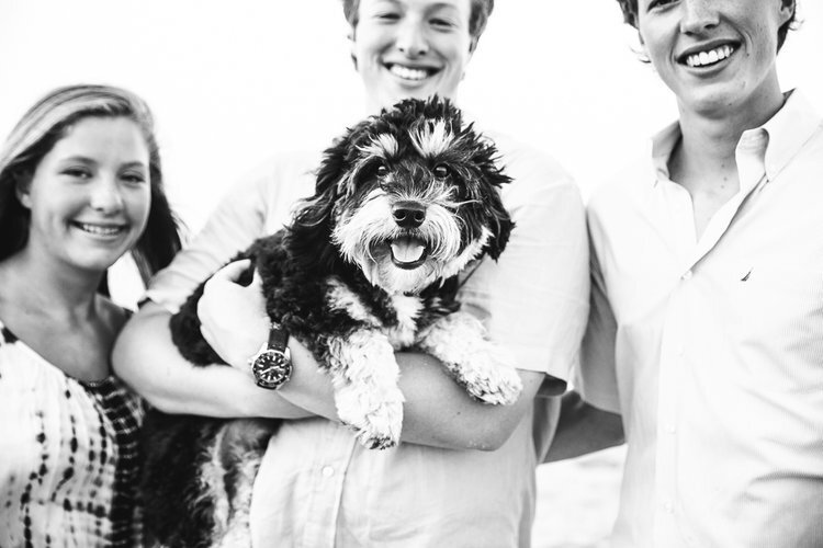  Black and white lifestyle photograph of two teen brothers and sister holding fluffy black and white dog smiling at camera in beach family photograph in Maryland. 