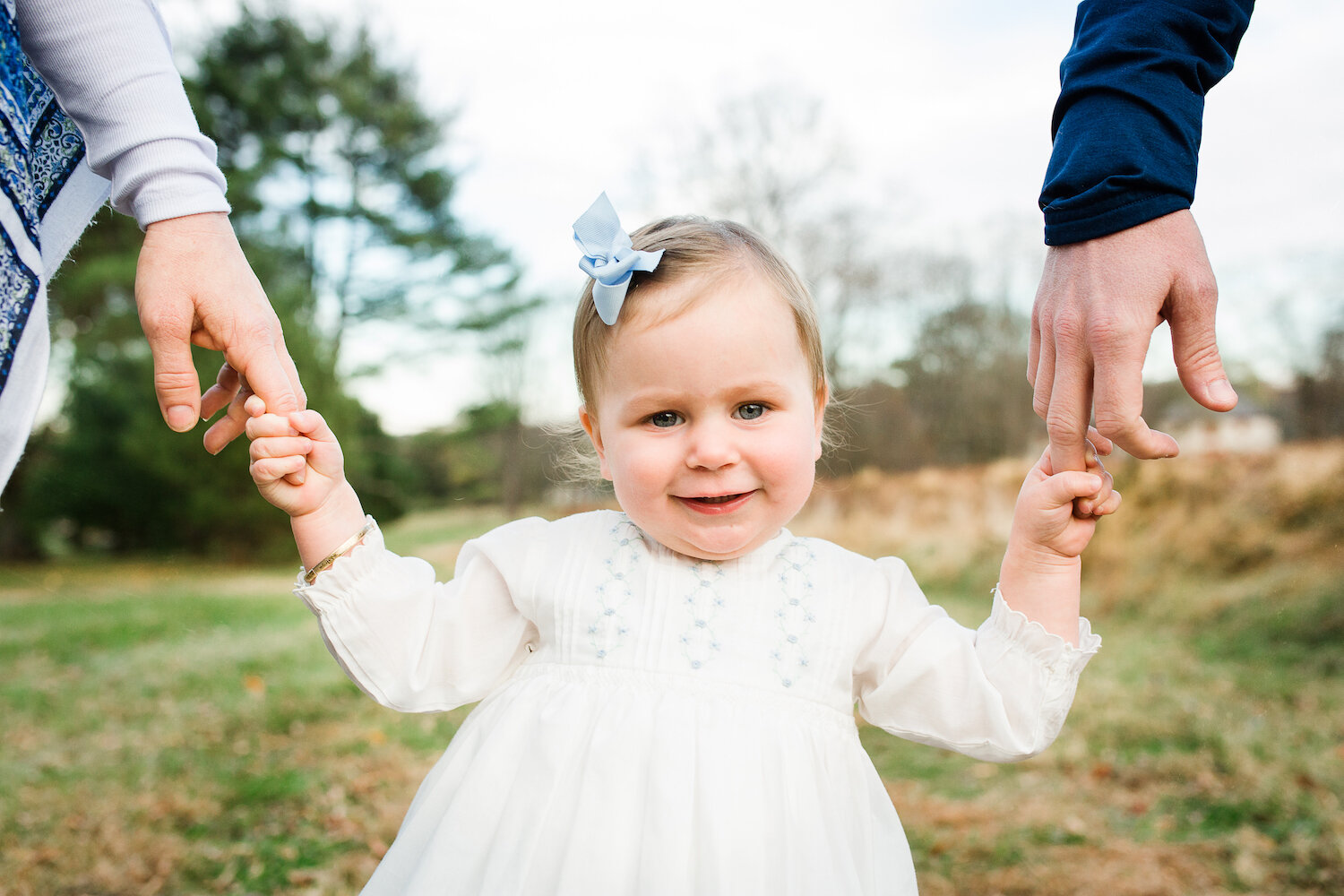 outdoor photograph of one year old girl with blonde hair and blue eyes holding parents hands