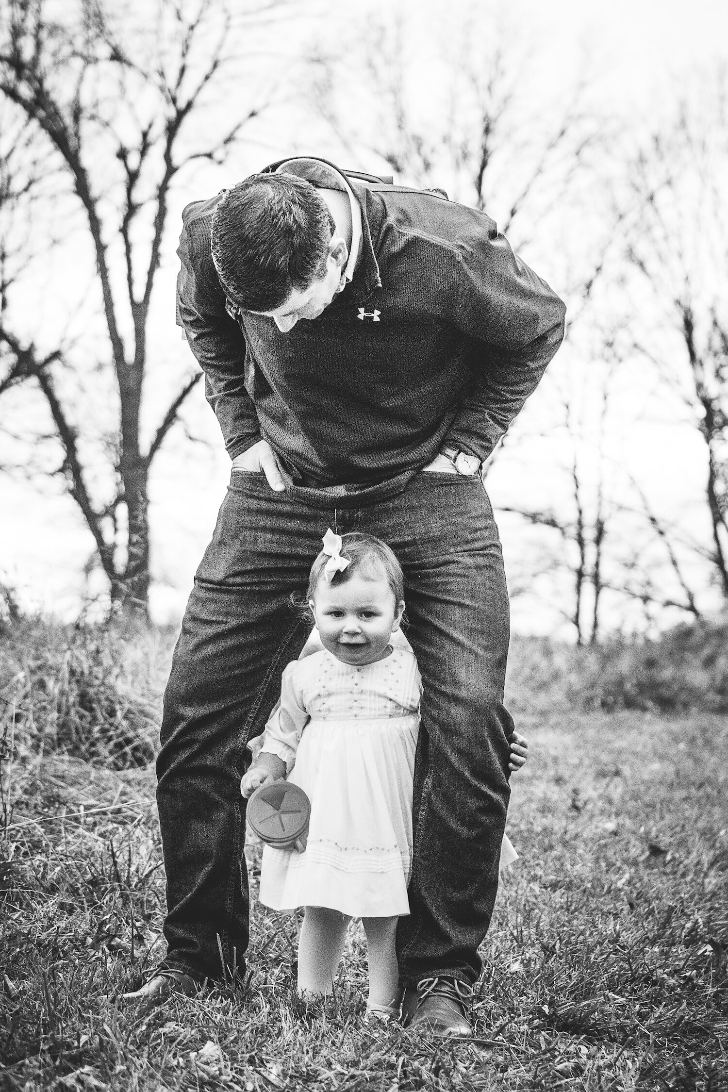 one year old little girl peeks through fathers legs in black and white family photo