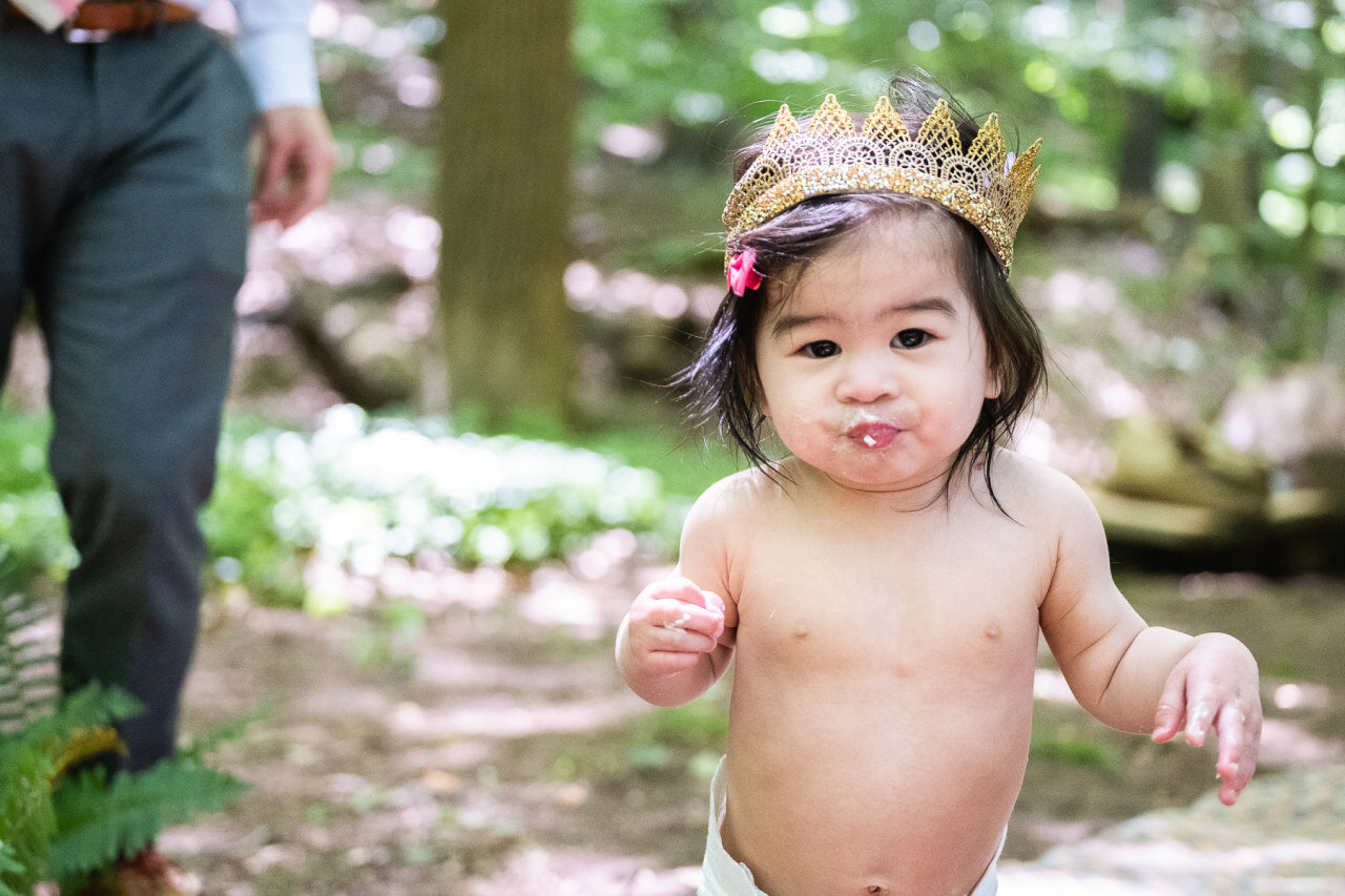one-year-old-girl-wearing-crown-eating-cake-smiling-at-camera.jpg