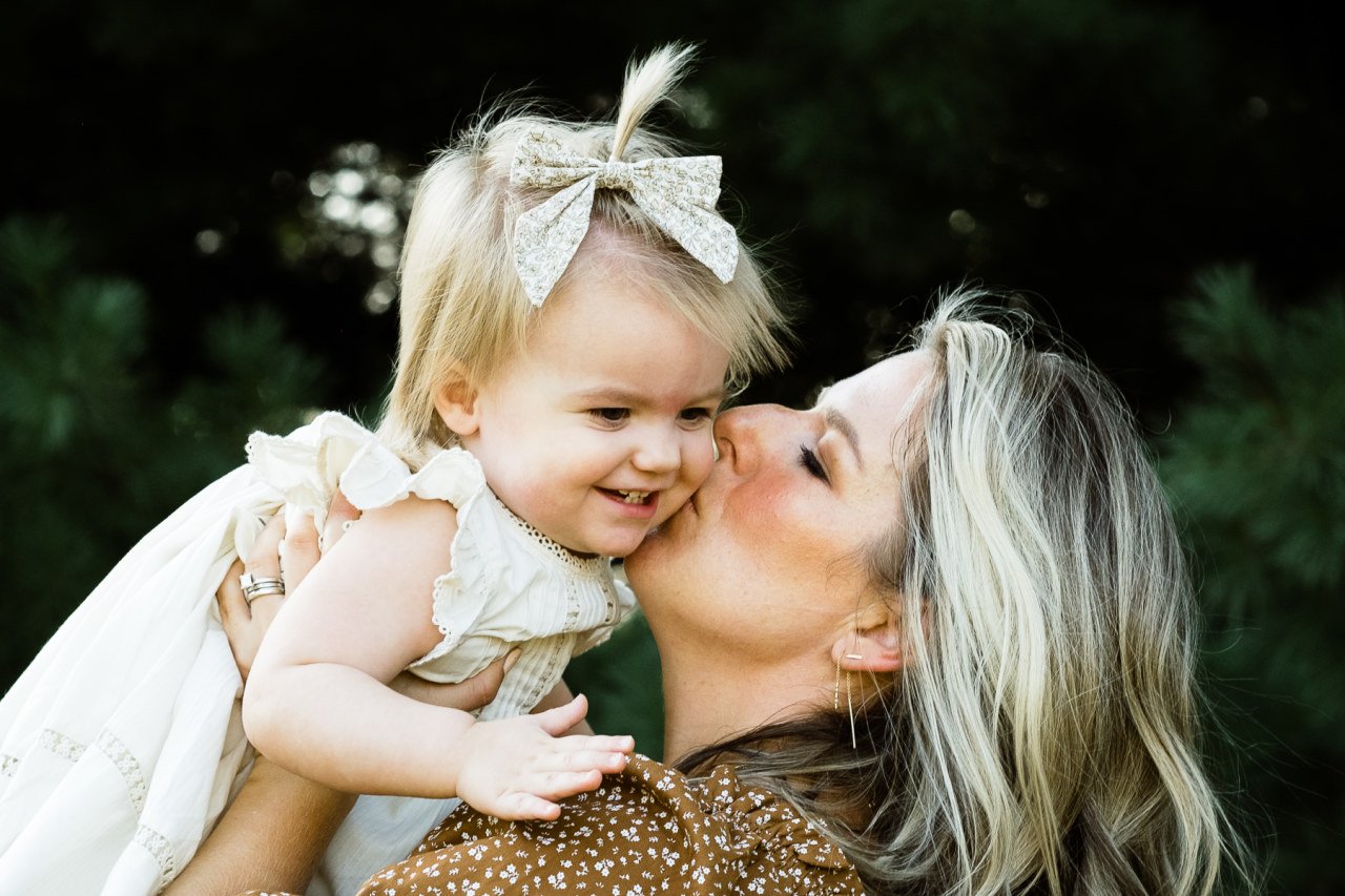 mother holds one year old daughter and kisses her on the cheek in family photograph