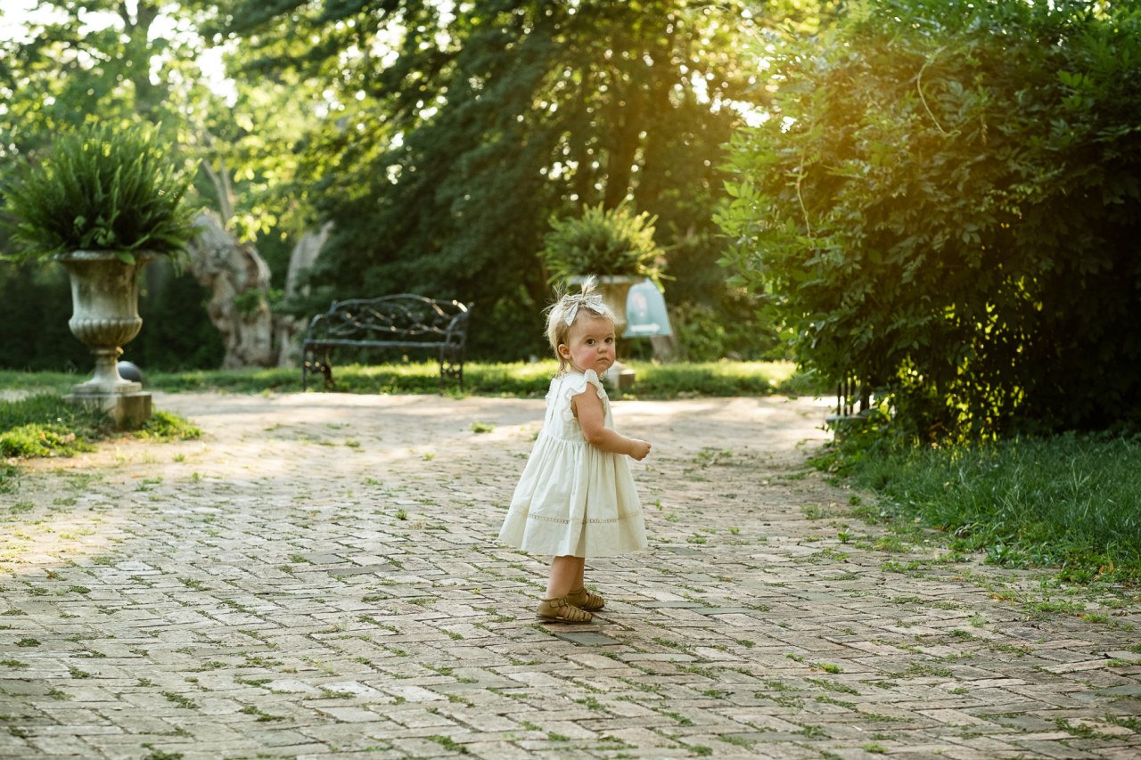 sweet toddler girl ables along brick path with sunlight glowing behind her.
