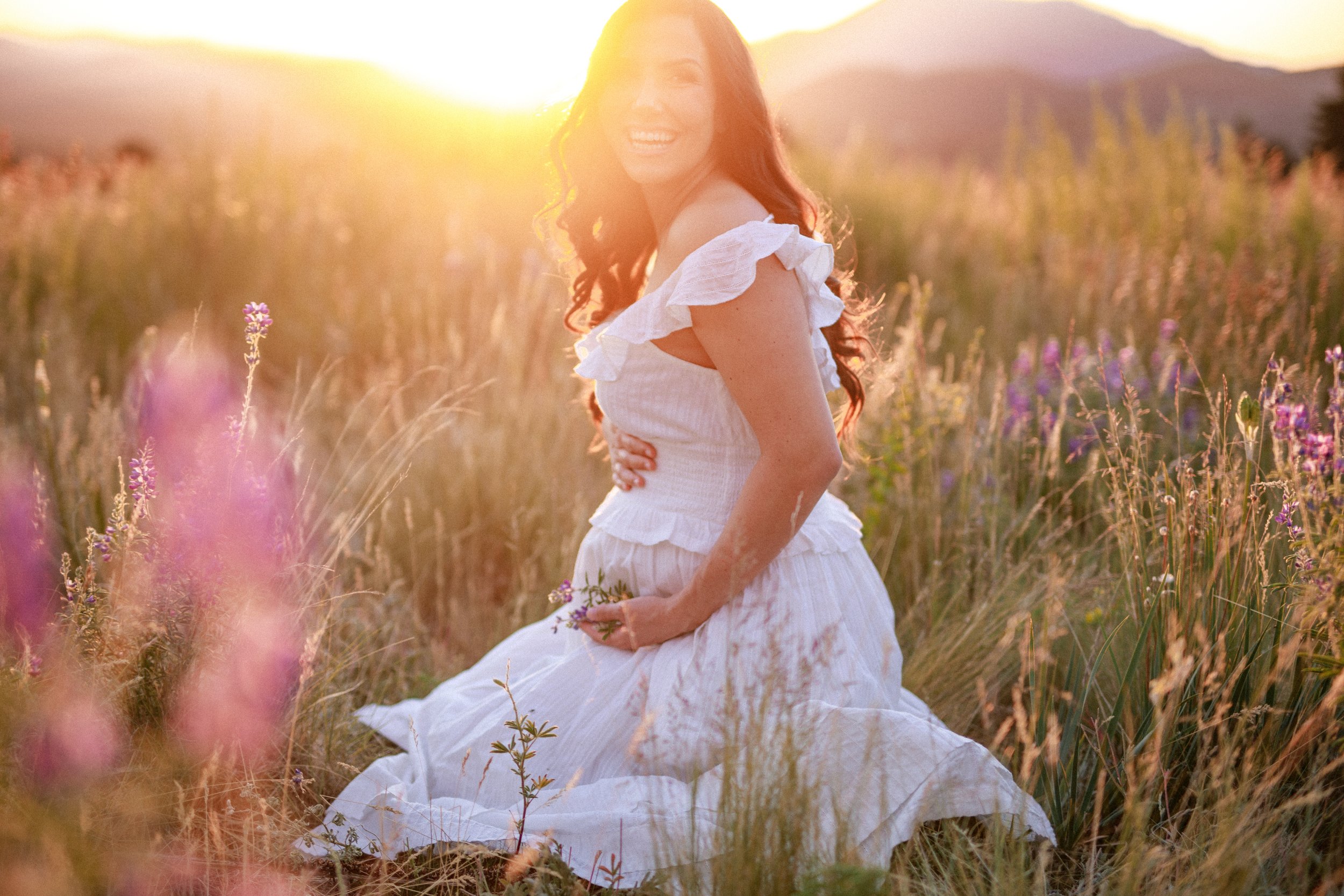 A woman in a white dress sitting in a field of flowers during sunset, smiling and holding flowers.